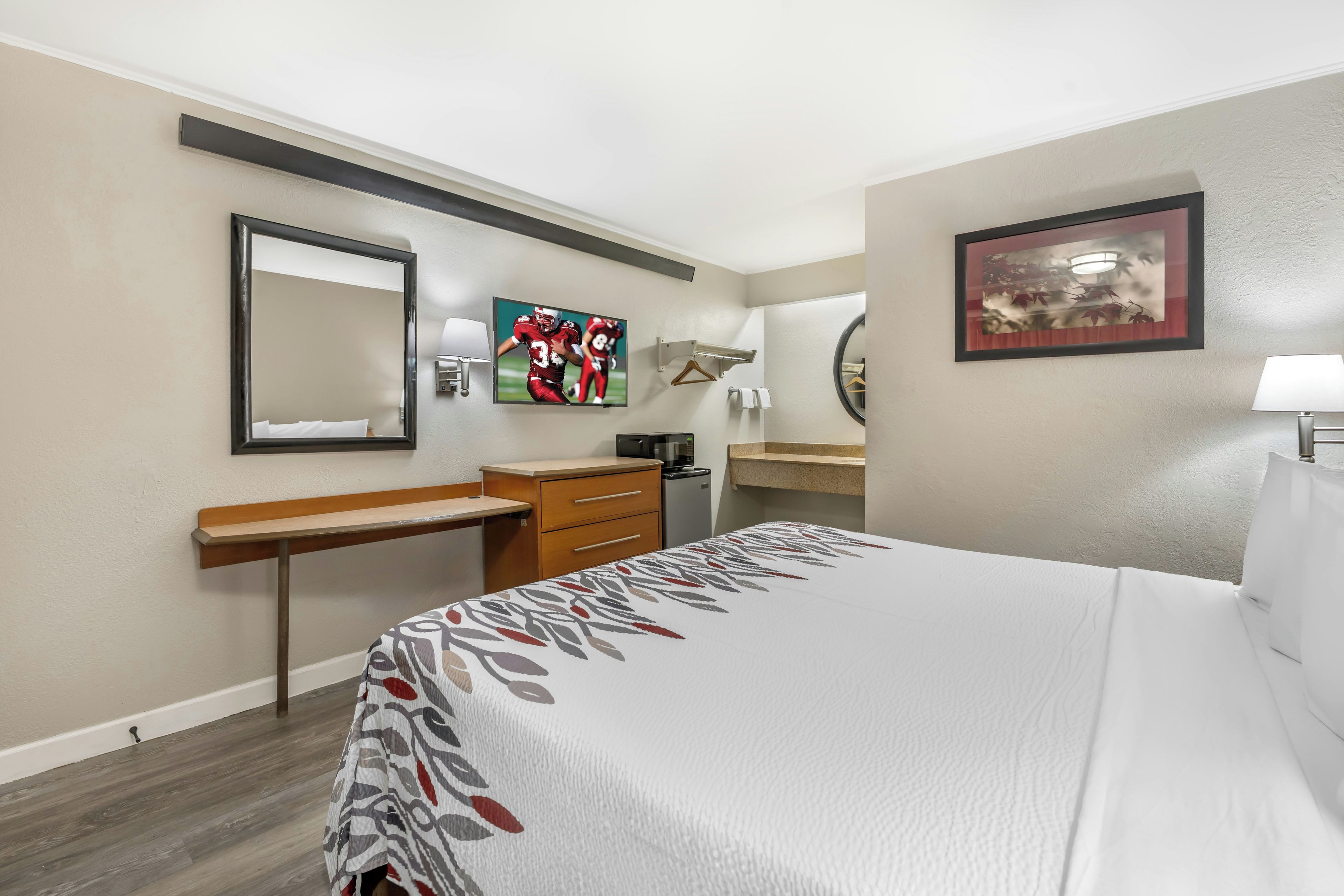 A hotel room with a bed, a desk, a chair, a mirror, a TV, and a bathroom.