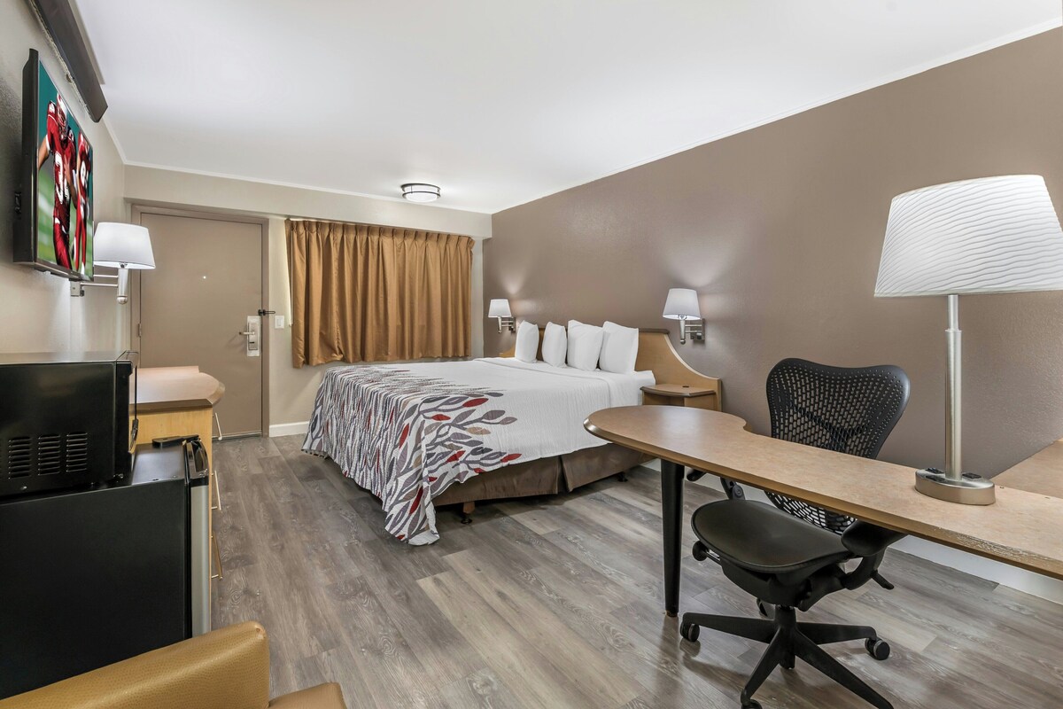 Quarto de hotel com cama, escrivaninha, cadeira, televisão e luminárias.