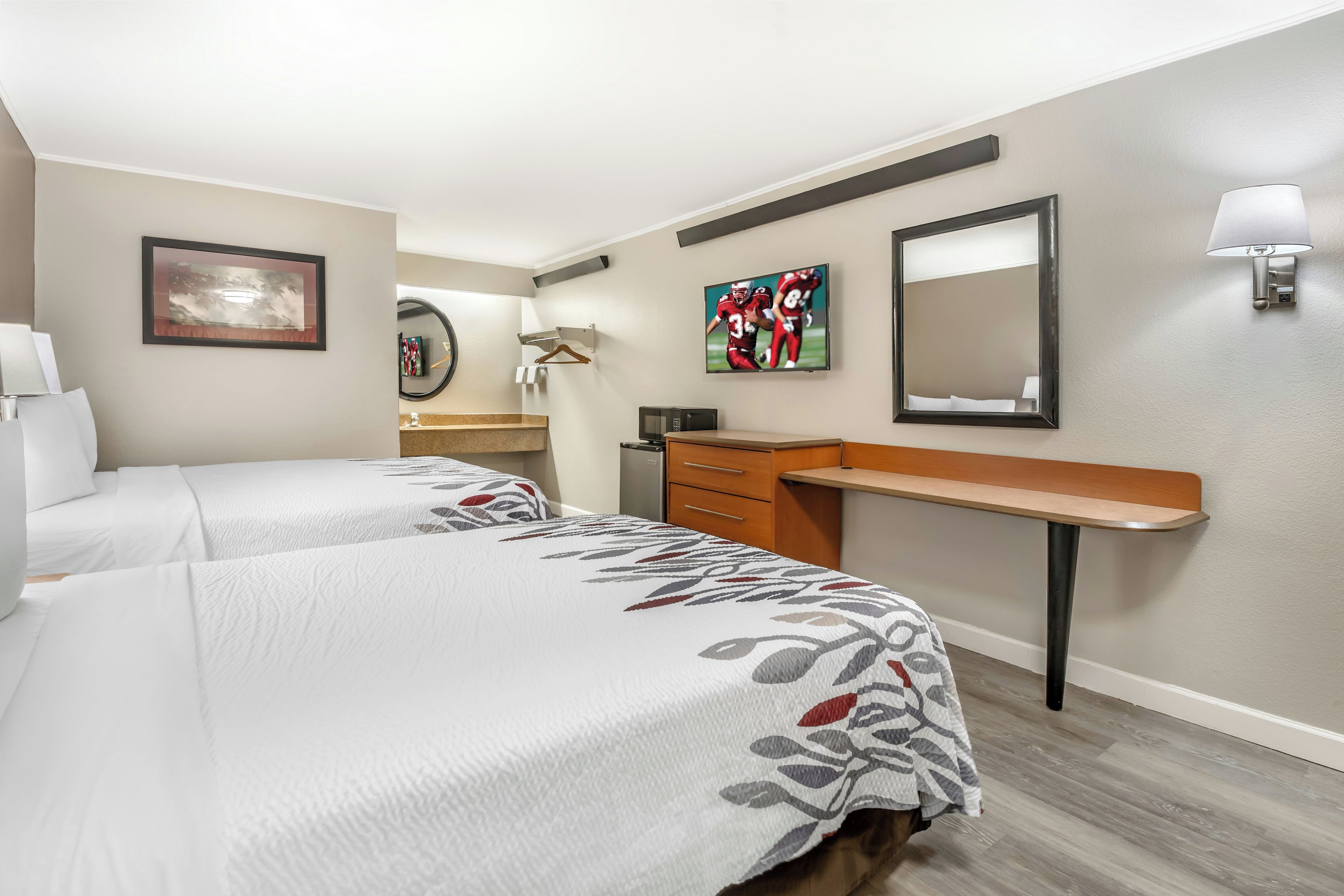 A hotel room with two beds, a desk, a mirror, a TV, and a lamp.