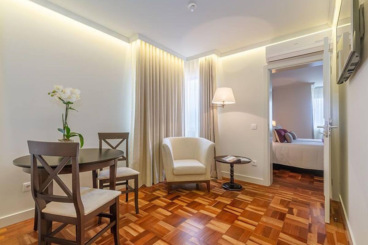 Une chambre d’hôtel avec un parquet en bois, une table à manger avec des chaises, une petite table ronde avec une lampe, un canapé et un lit avec des oreillers.
