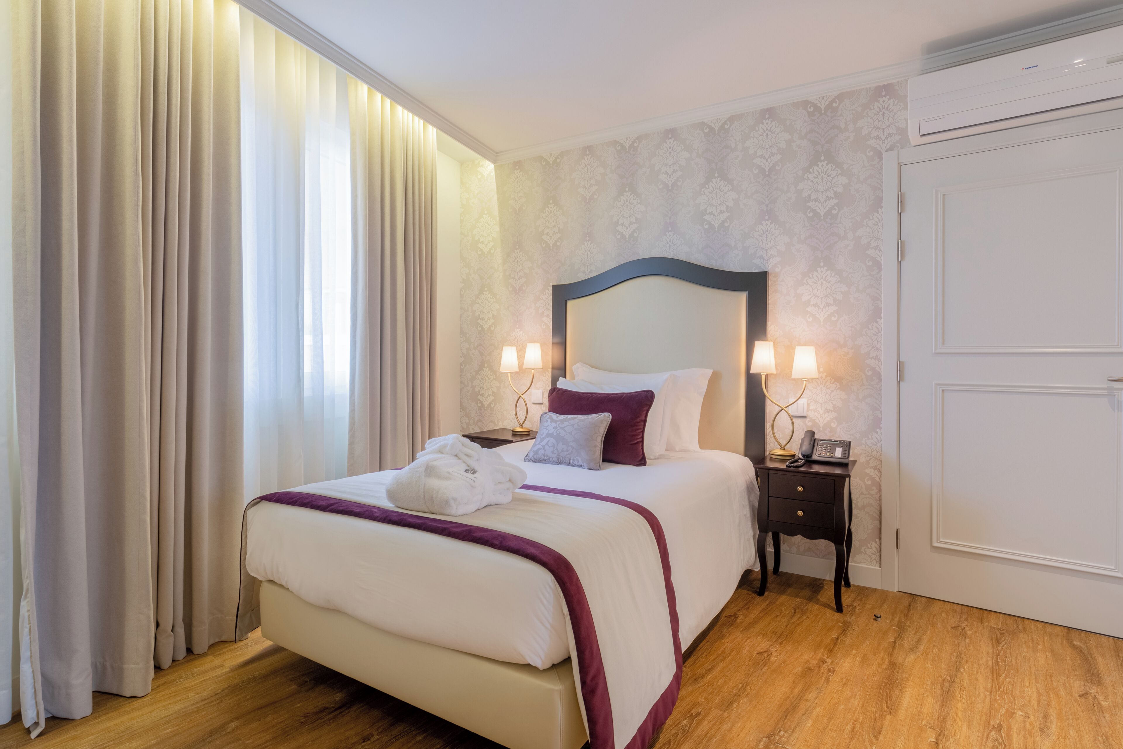 A hotel room with a bed, curtains, a nightstand, and a wall-mounted air conditioner.
