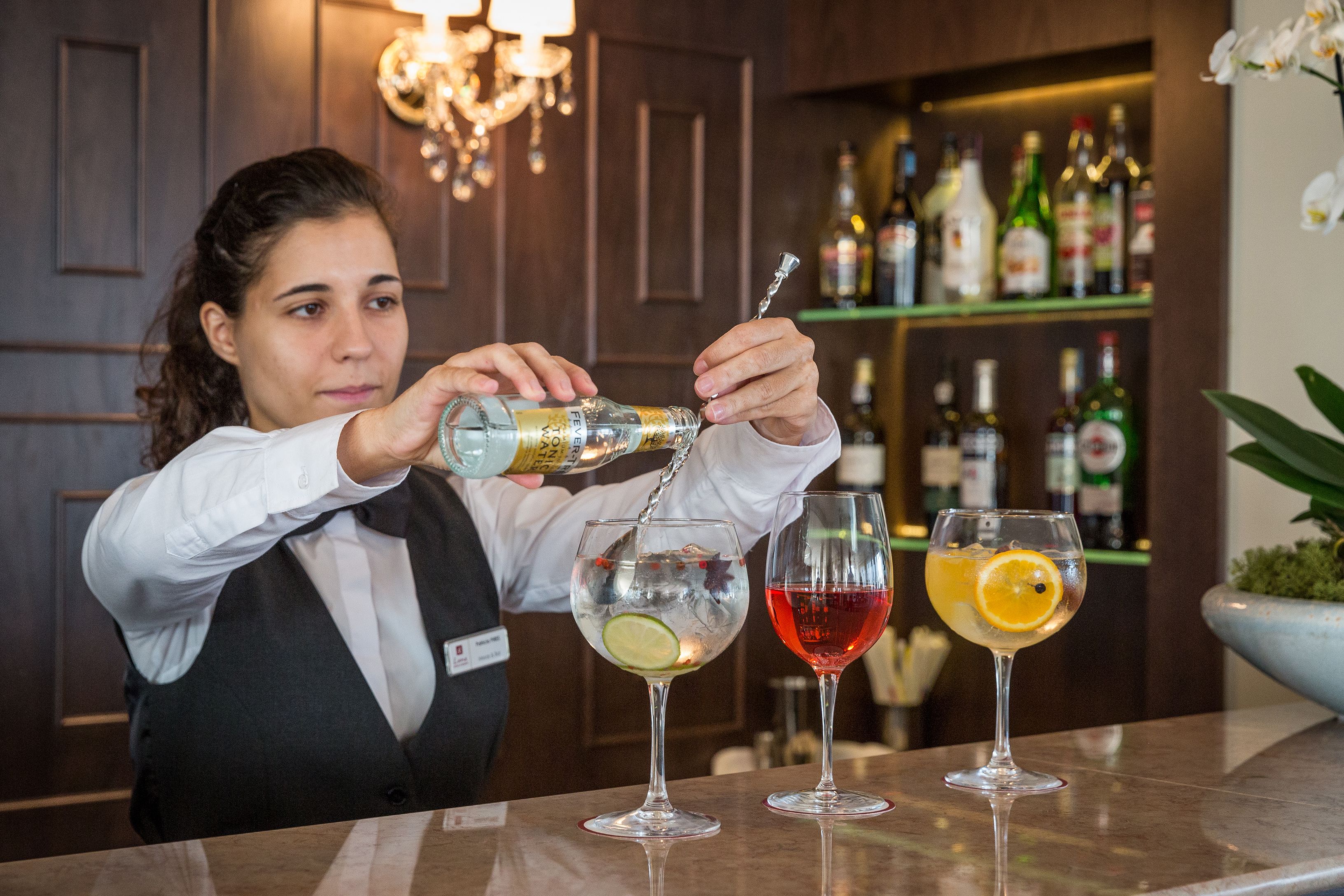 En bartender som blandar drinkar vid en bar, med ett stort antal flaskor synliga i bakgrunden.