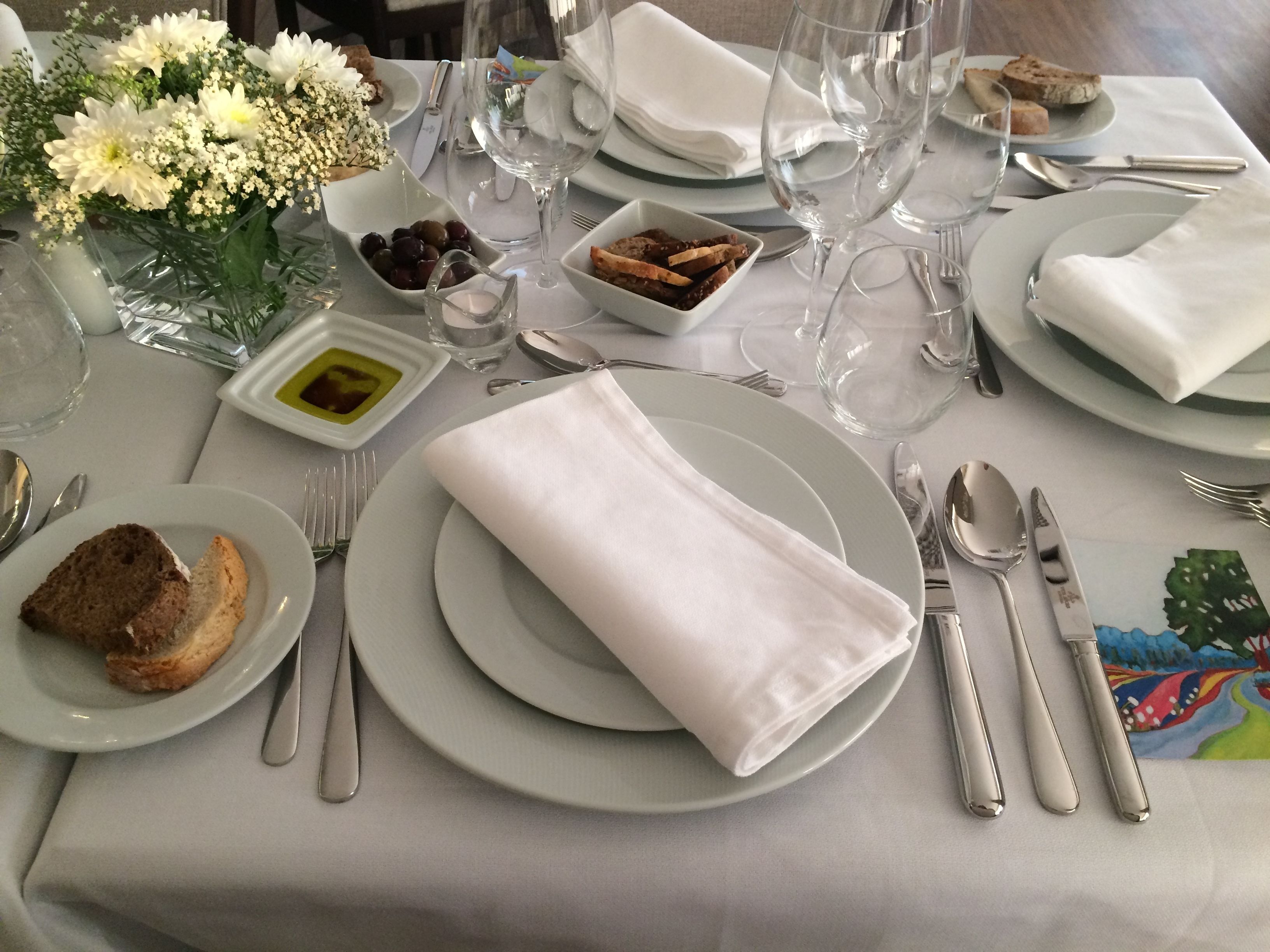 Una mesa dispuesta para una comida, con platos, cubiertos, vasos y un pequeño jarrón con flores.