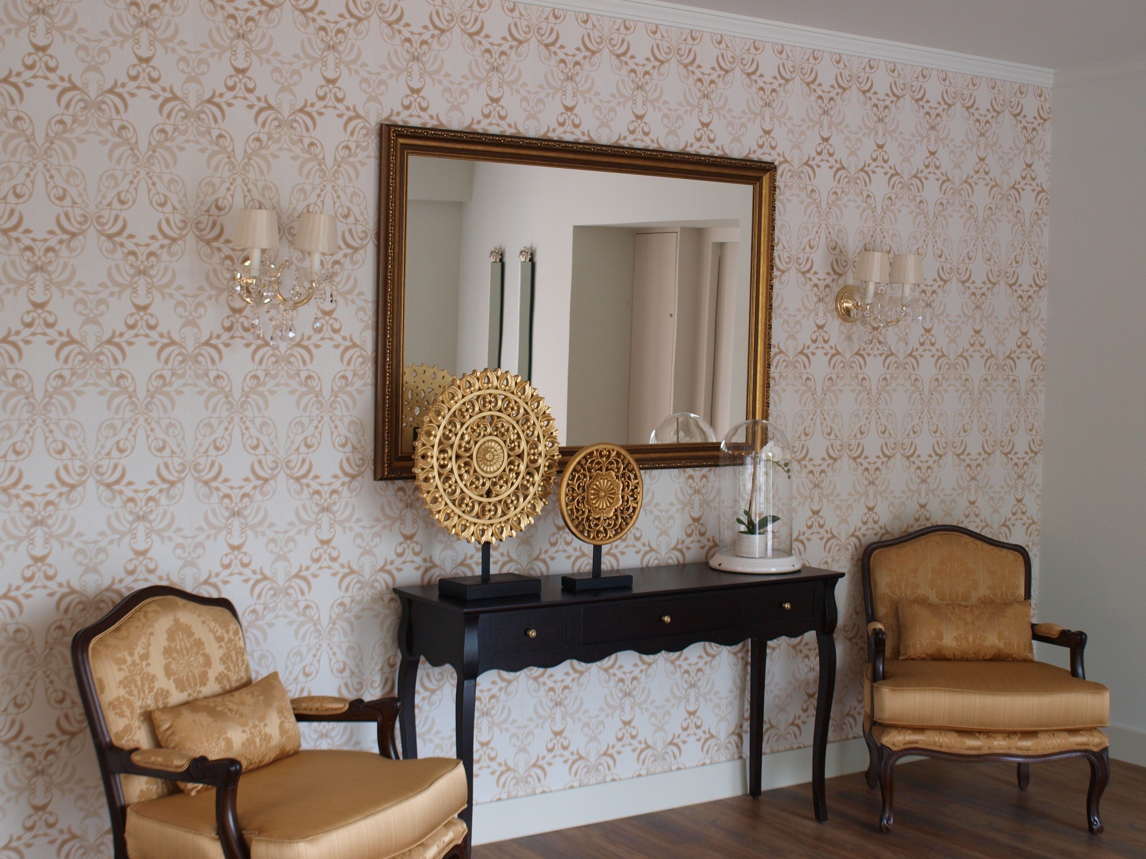 A room with patterned wallpaper, a large mirror, two chairs, a console table with decorative items, and wall sconces.