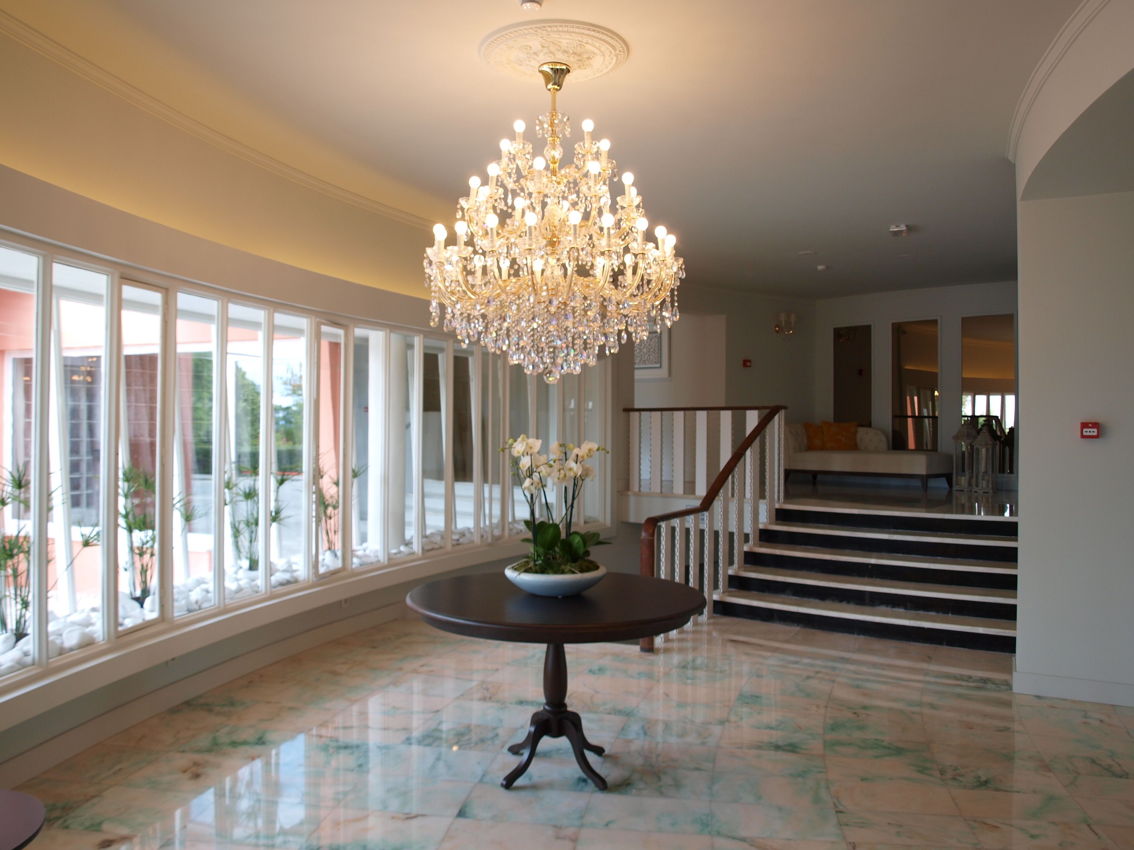 Um amplo hall com um lustre imponente, uma mesa redonda com um vaso de flores e uma escadaria que dá acesso aos andares superiores.