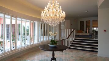 Um amplo hall com um lustre imponente, uma mesa redonda com um vaso de flores e uma escadaria que dá acesso aos andares superiores.
