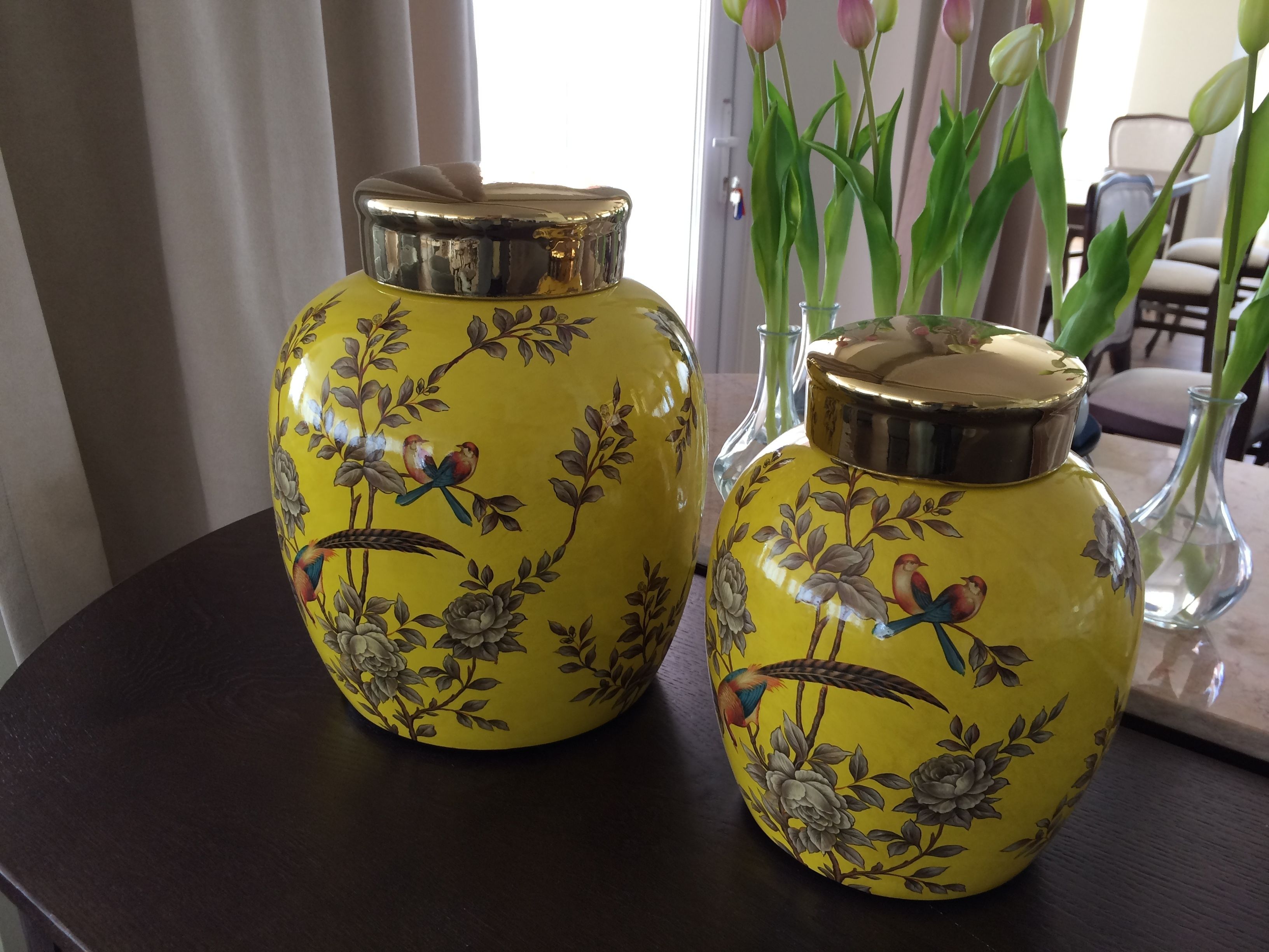 Two yellow ceramic vases with metallic lids, featuring bird and floral designs, placed on a dark wooden table.