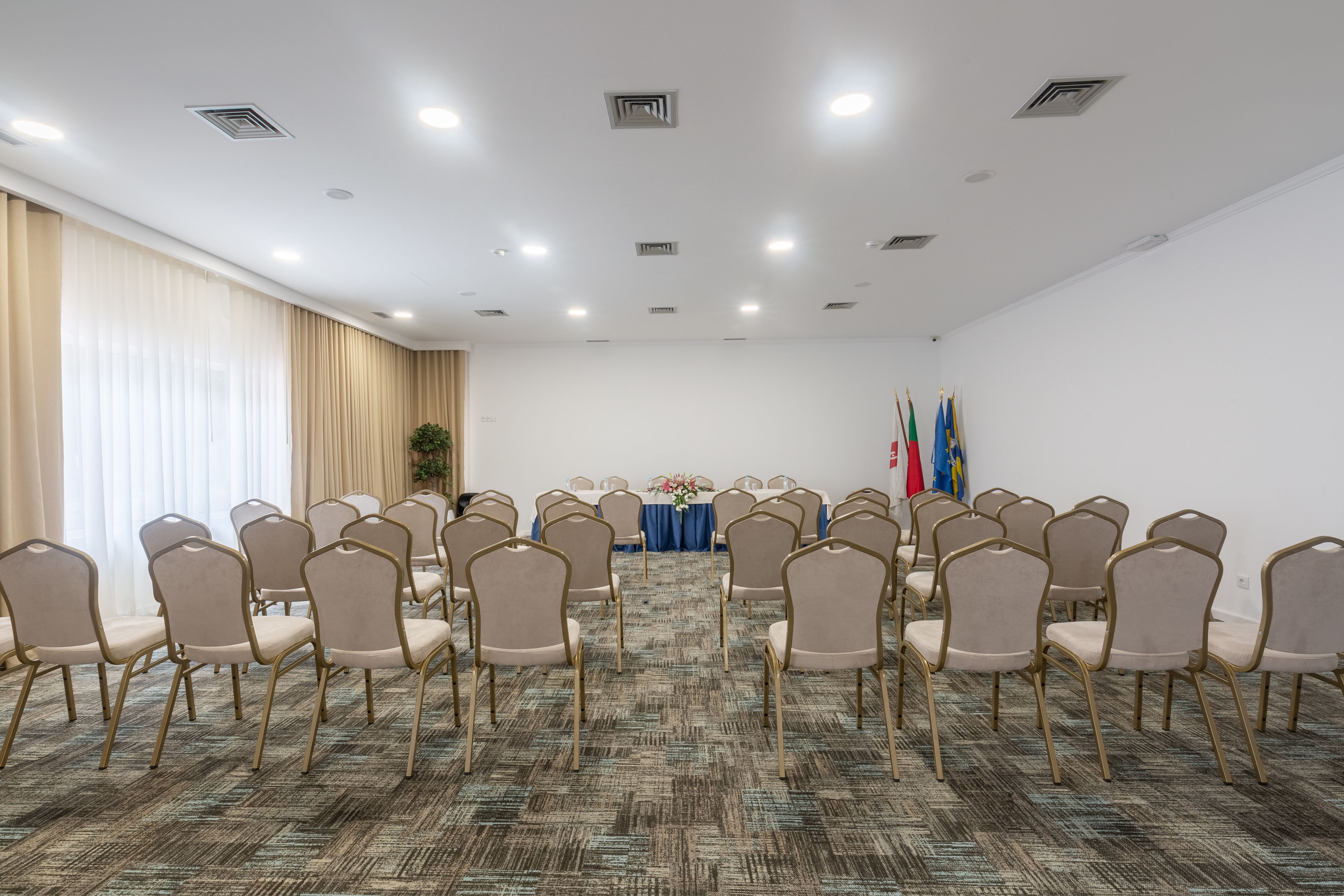 A conference room with rows of chairs, a table with a blue tablecloth, flags, and a patterned carpet.