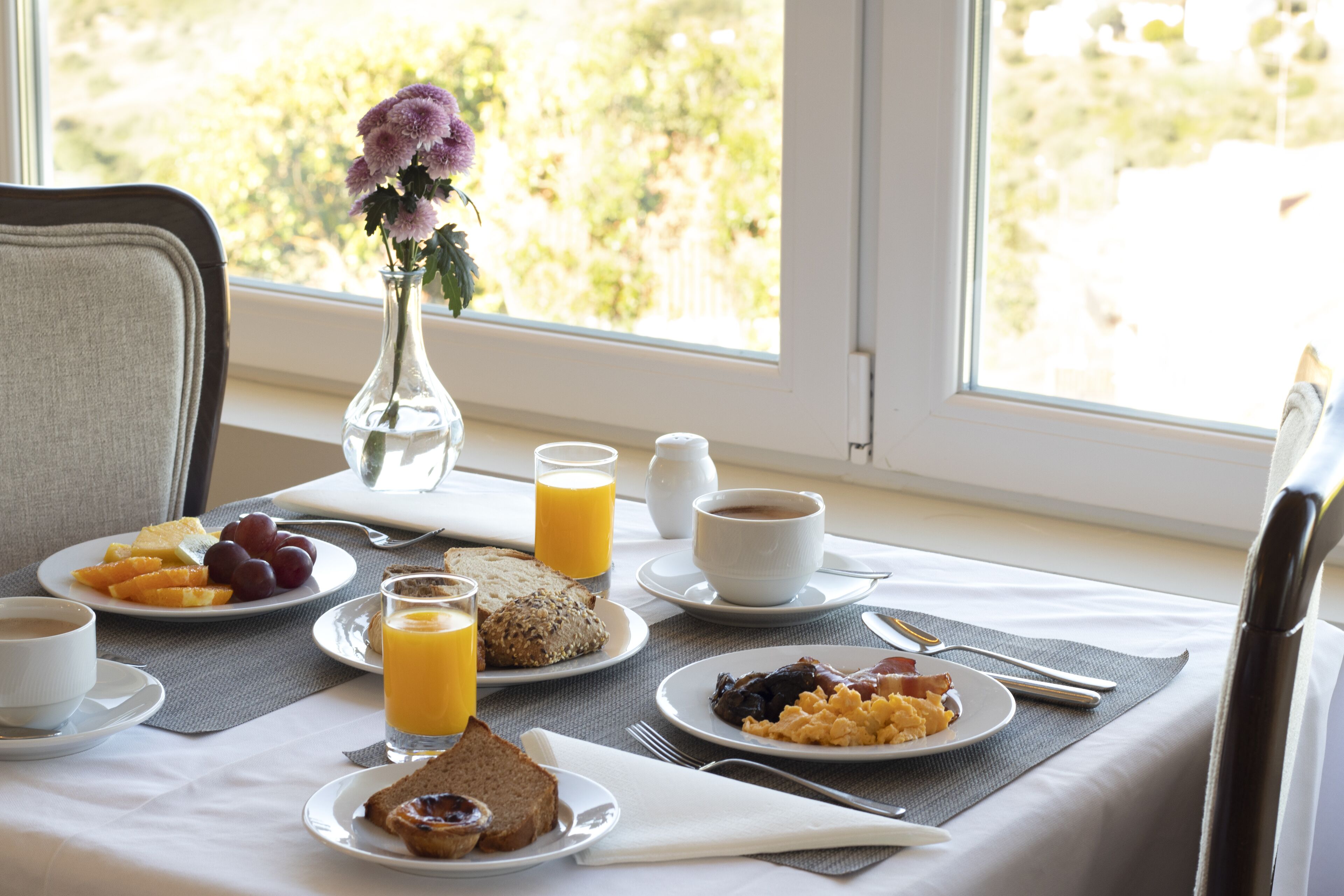 Une table de petit-déjeuner avec des assiettes remplies de nourriture, un vase de fleurs et une fenêtre donnant sur des arbres.