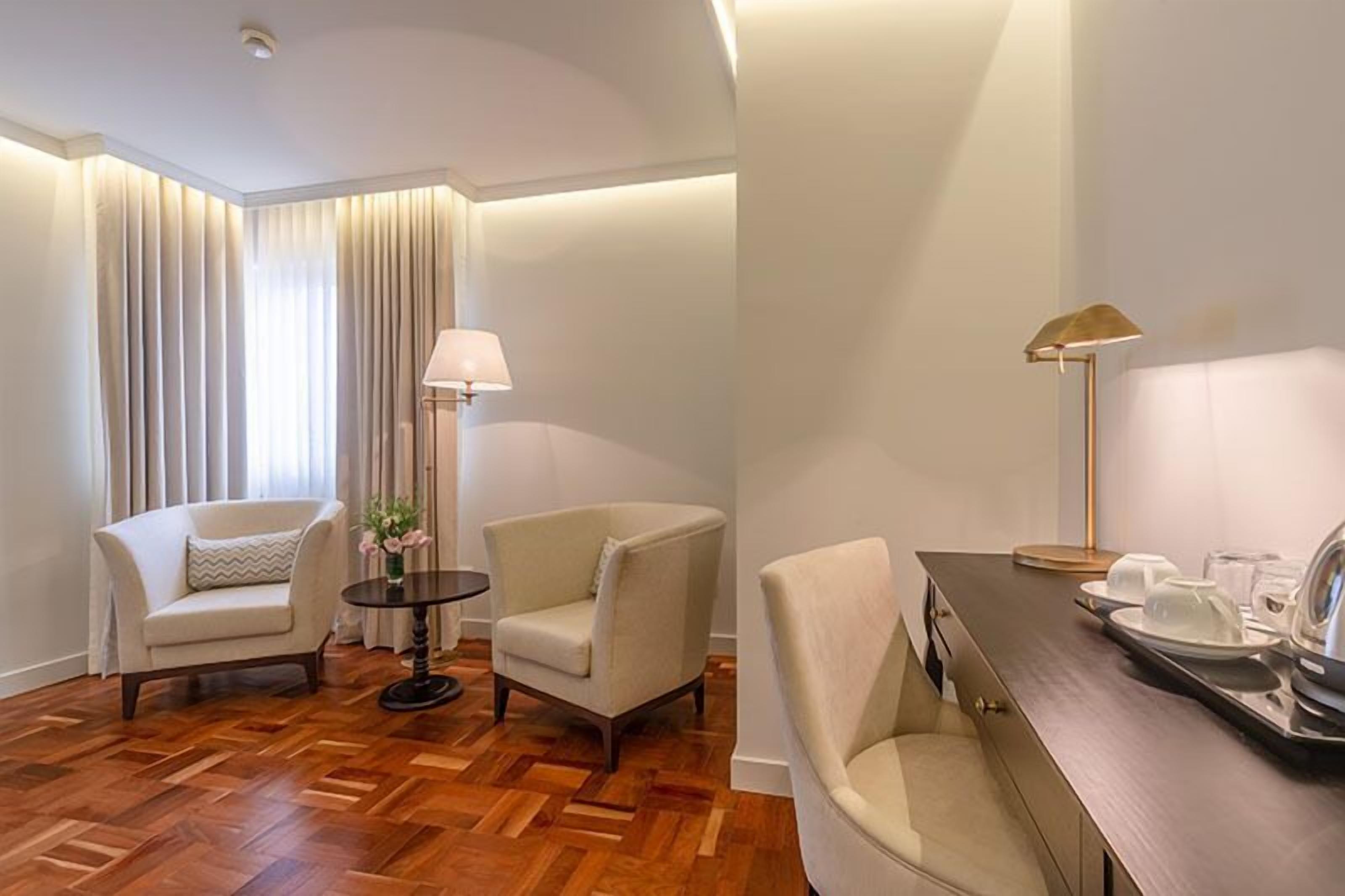 A hotel room with a wooden parquet floor, two armchairs, a round table with a lamp, and a dining table set with plates and cups.