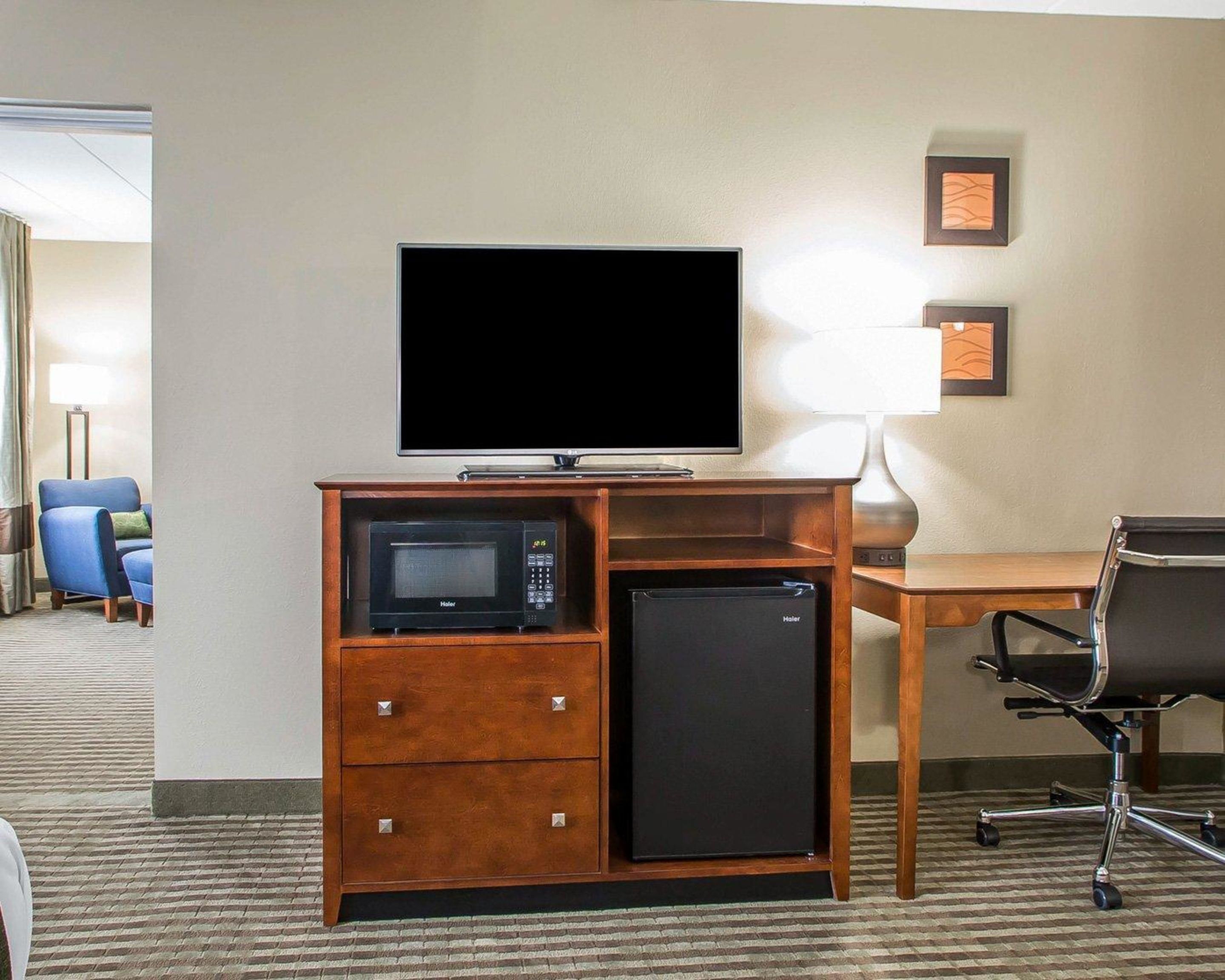 A hotel room with a flat-screen TV on a wooden stand, a microwave, a mini fridge, a desk, and a chair.