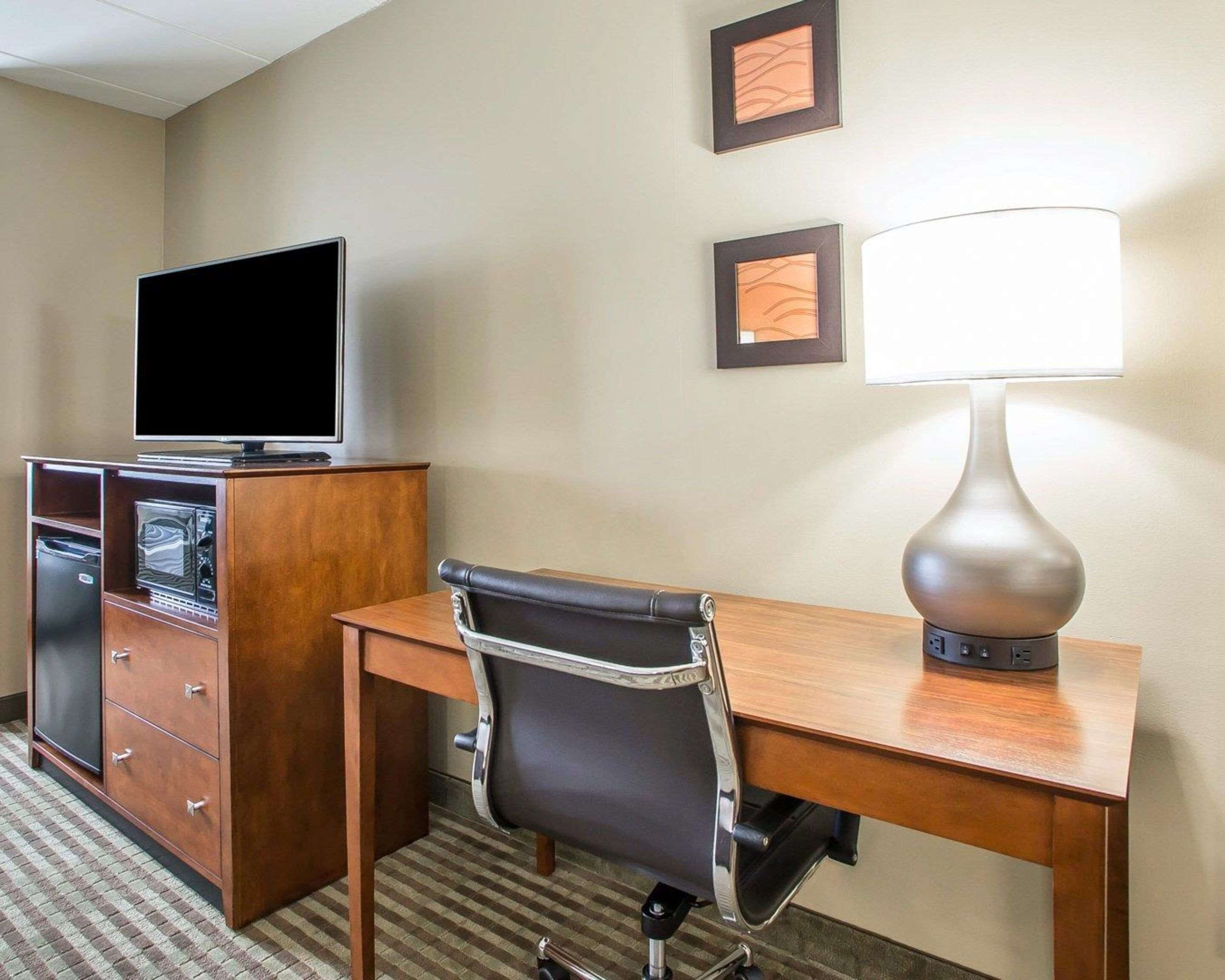 A hotel room with a wooden desk, a black office chair, a flat-screen TV on a cabinet, a lamp, and two framed pictures on the wall.