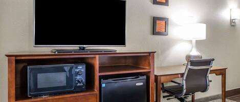 Habitación de hotel con televisión de pantalla plana, microondas, refrigerador pequeño, escritorio, silla y lámpara.