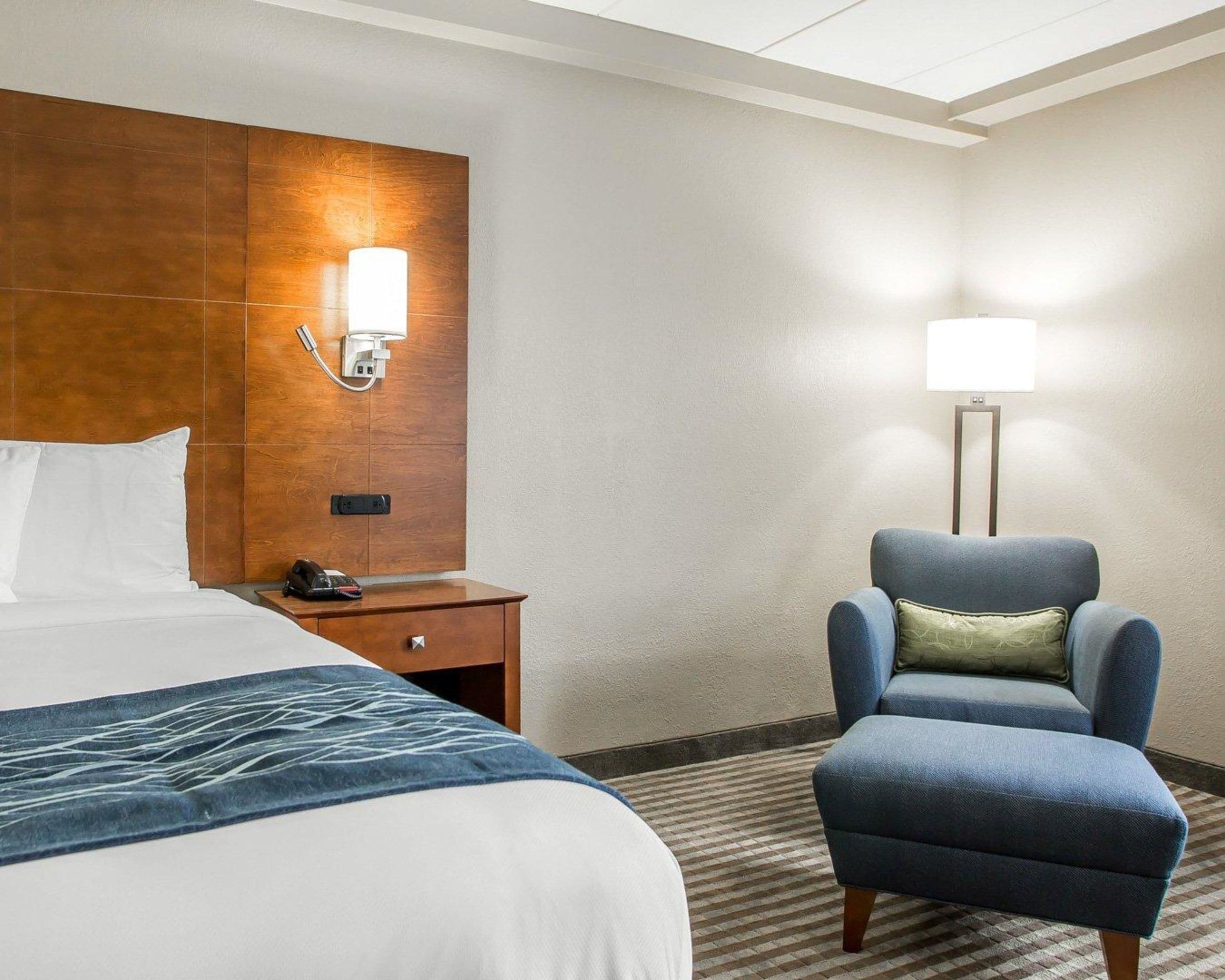 Une chambre d’hôtel avec un grand lit, une tête de lit en bois, un bureau avec un téléphone, un fauteuil bleu et une lampe sur pied.