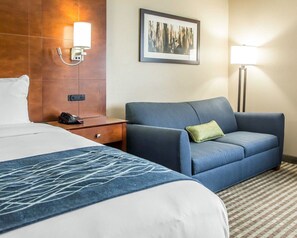 A hotel room with a bed, a blue sofa, a nightstand with a phone, a wall-mounted light, and a framed picture. - Comfort Inn Hammond I-94 (Hammond)