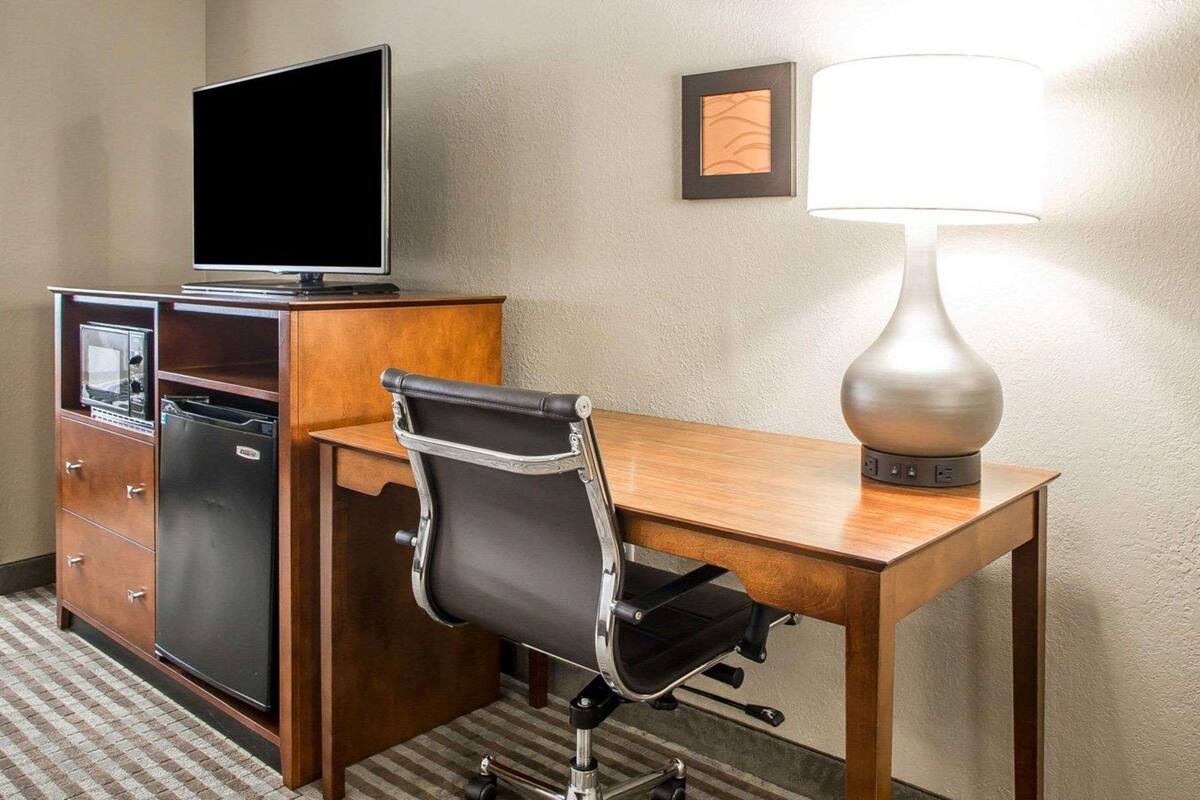 A hotel room with a wooden desk, a black office chair, a television on a cabinet, and a wall-mounted lamp.