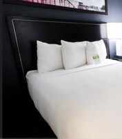 Habitación de hotel con cabecera negra, ropa de cama blanca y una pequeña etiqueta verde en la almohada.