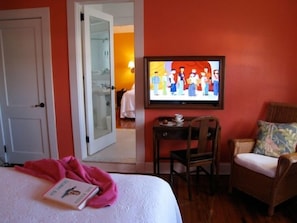 Quarto de hotel com cama, televisão na parede, cadeira e pequena mesa com xícara e pires.