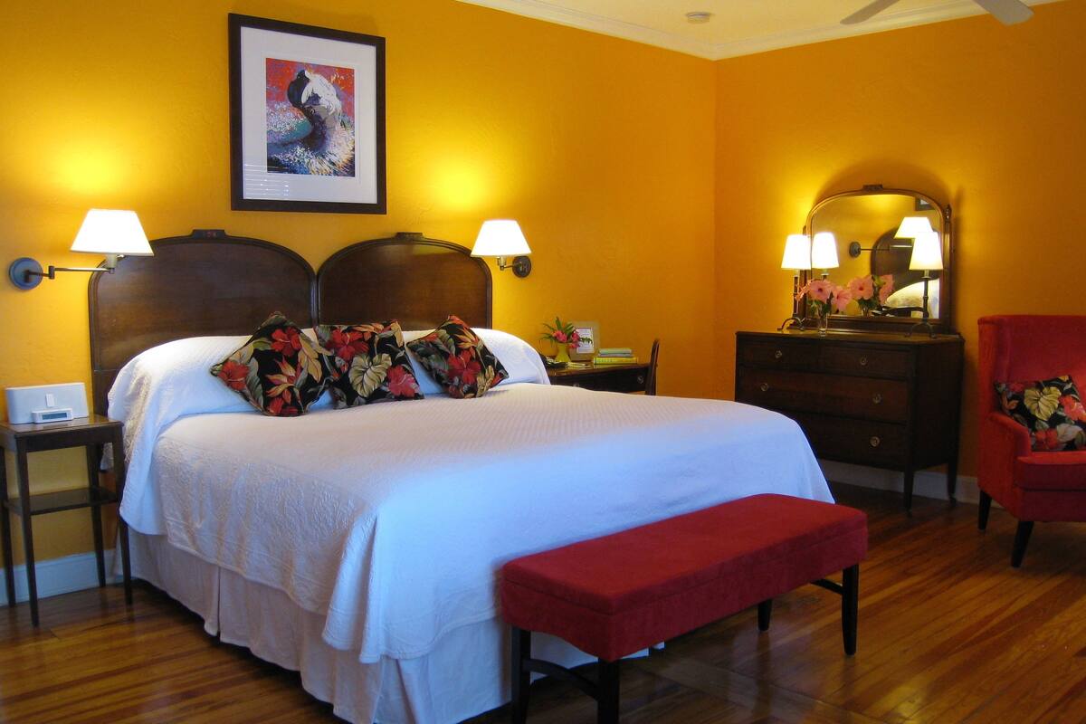 A hotel room with a large bed, two bedside lamps, a wooden headboard, a red armchair, a wooden dresser, and a framed picture on the wall.