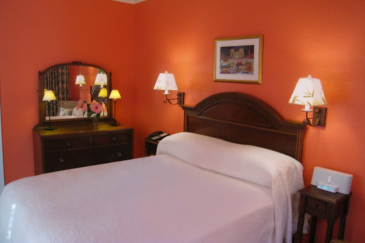 A hotel room with a large bed, a dresser with a mirror, two wall-mounted lamps, a bedside table with a lamp, and a framed picture on the wall.