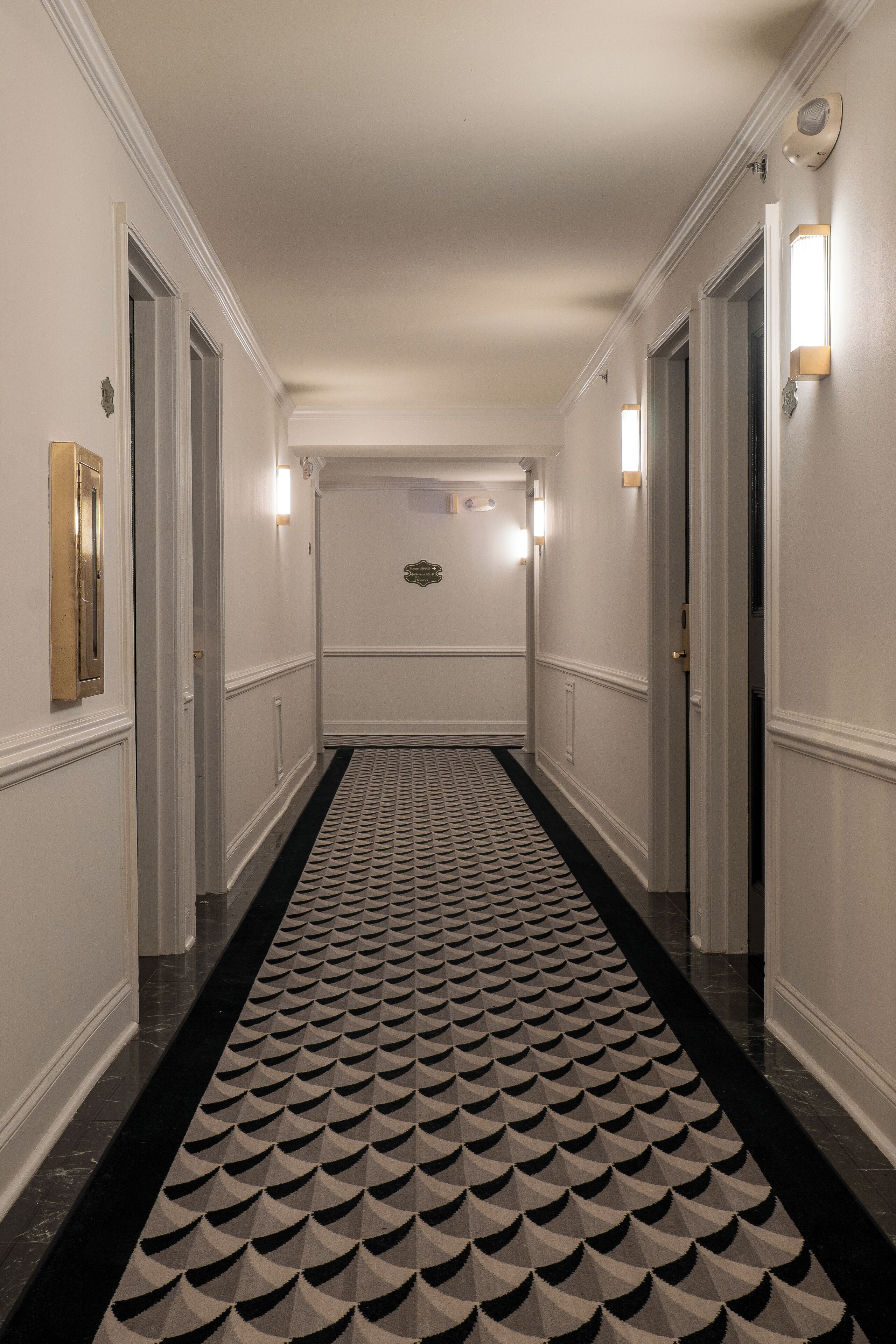Un couloir aux murs blancs, avec un tapis à motifs et des portes de part et d’autre.