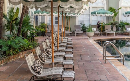 A row of wicker lounge chairs with green umbrellas, a pool, and a building with balconies in the background.