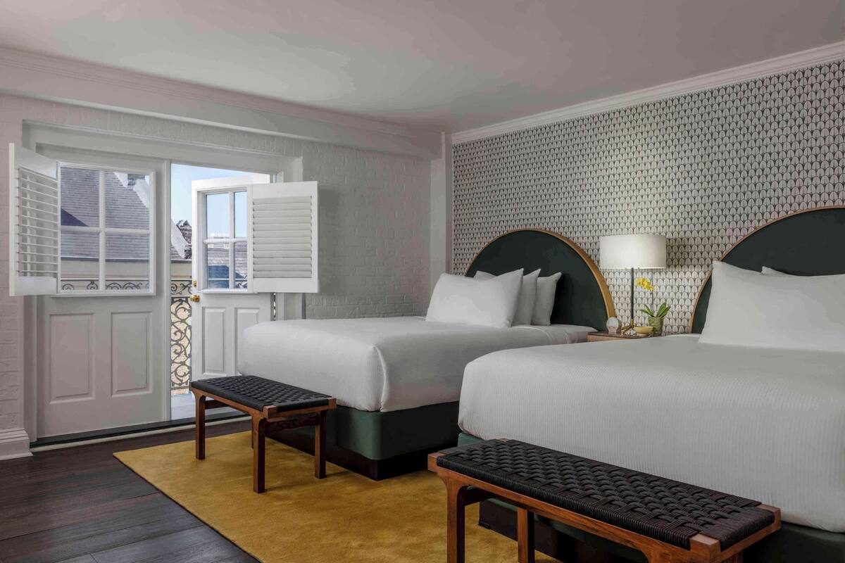 Een hotelkamer met twee bedden, een raam met zonwering, een nachtkastje met een lamp en een behang met een patroon.