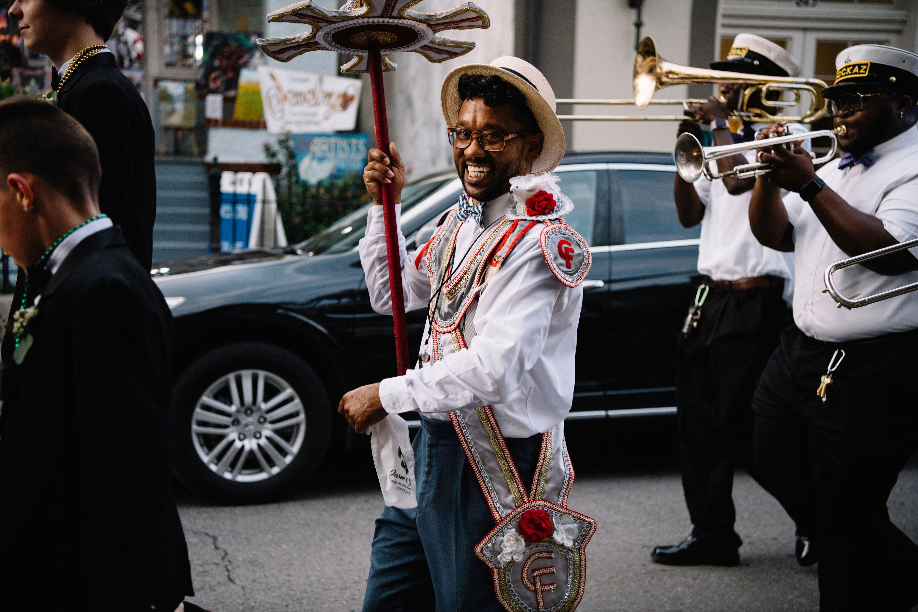 Una persona con camisa y sombrero blancos, sosteniendo un bastón rojo, con un lazo decorativo y una flor.