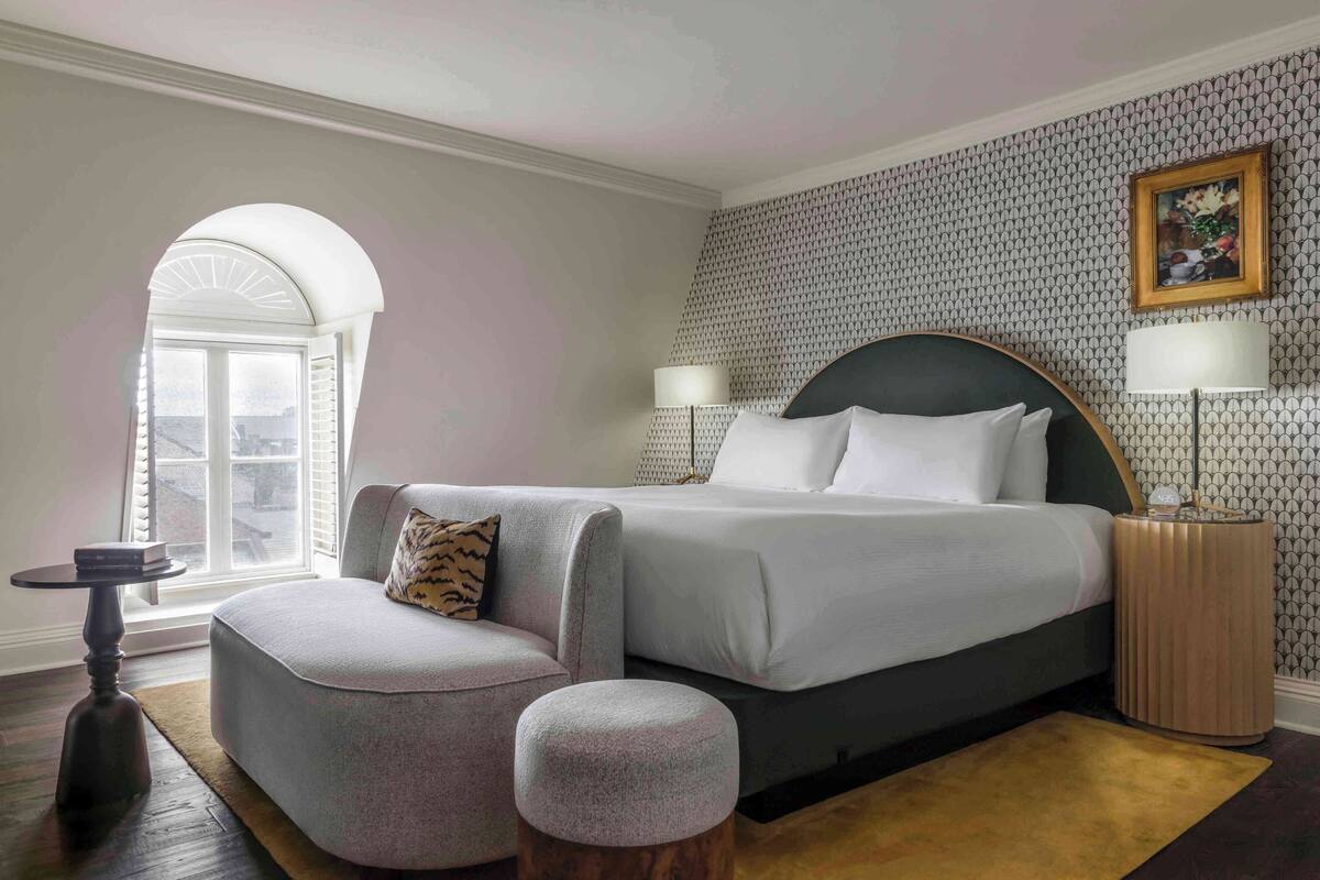 Een slaapkamer met een groot bed, een bank, een rond voetenbankje, een klein tafeltje en een raam met luiken.