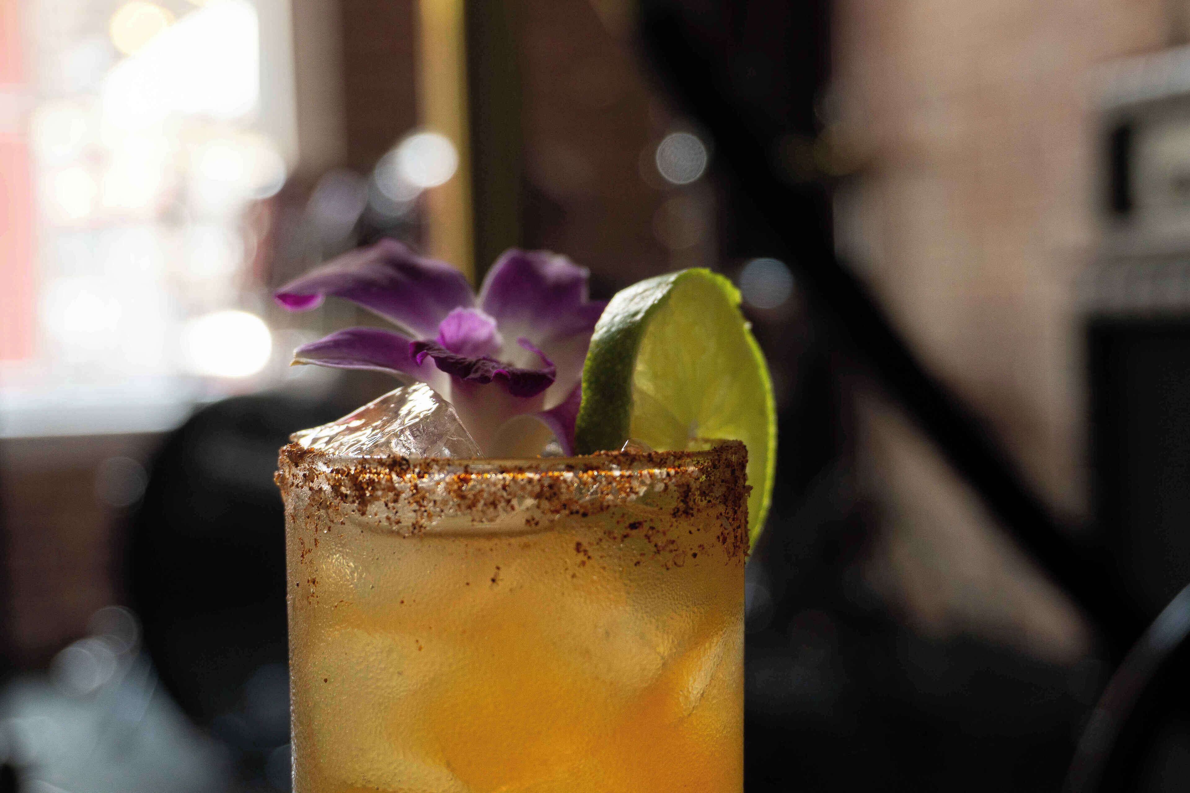 A close-up of a cocktail with a salted rim, lime wedge, and purple flower garnish.