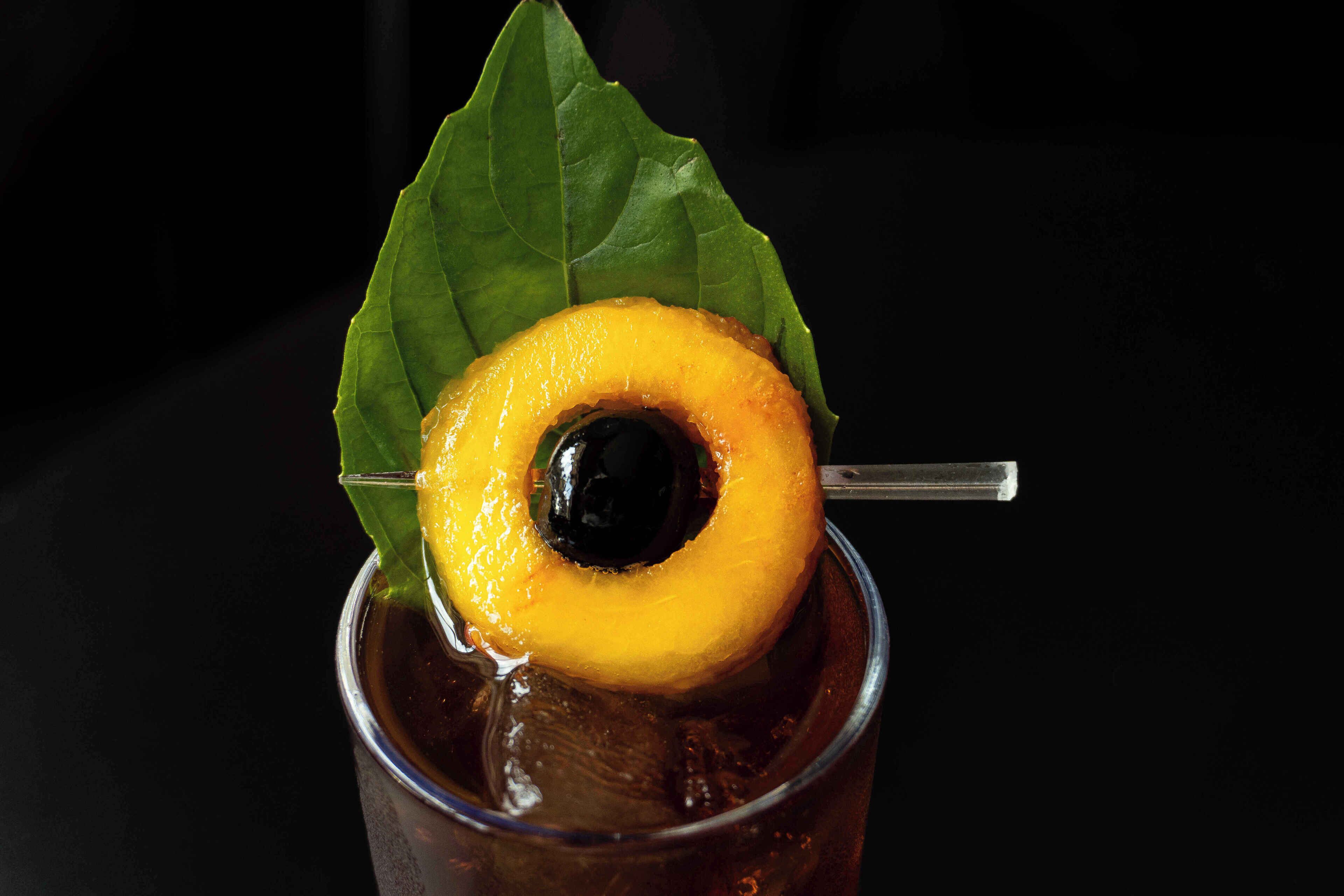 A glass with a dark liquid, a yellow fruit slice, and a green leaf.