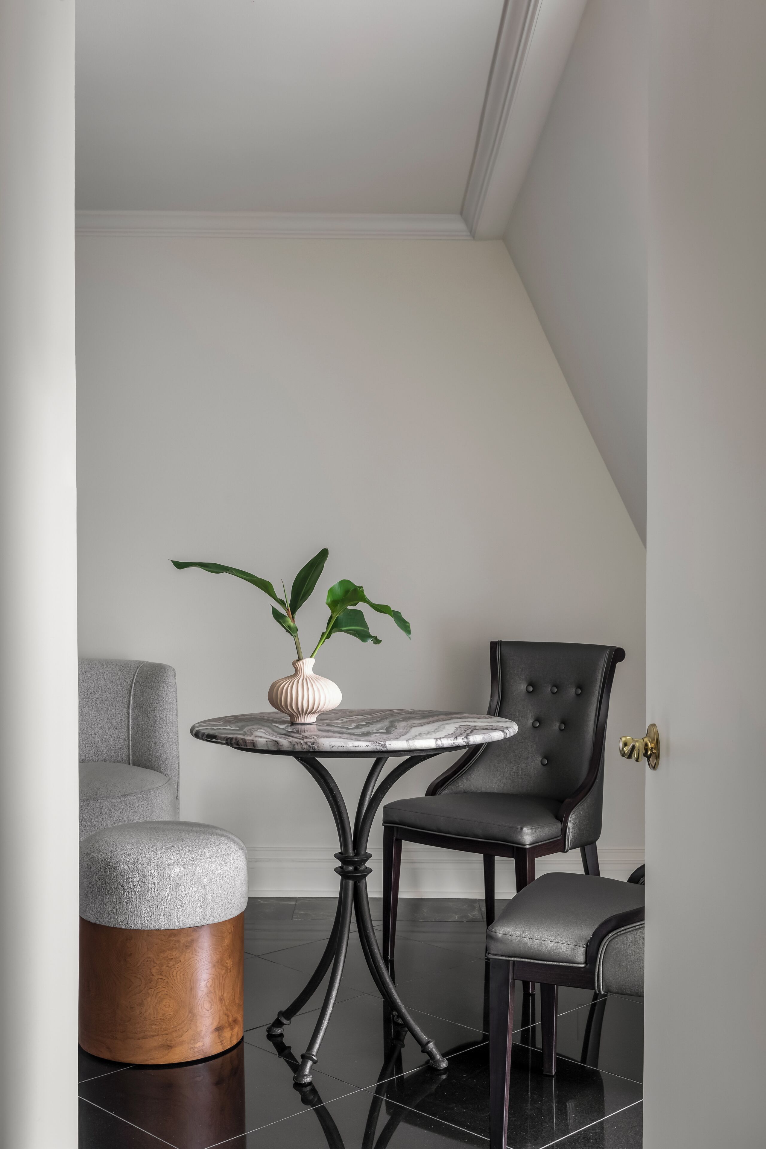 A corner room with a round table, a grey armchair, and a black chair with a decorative door handle.