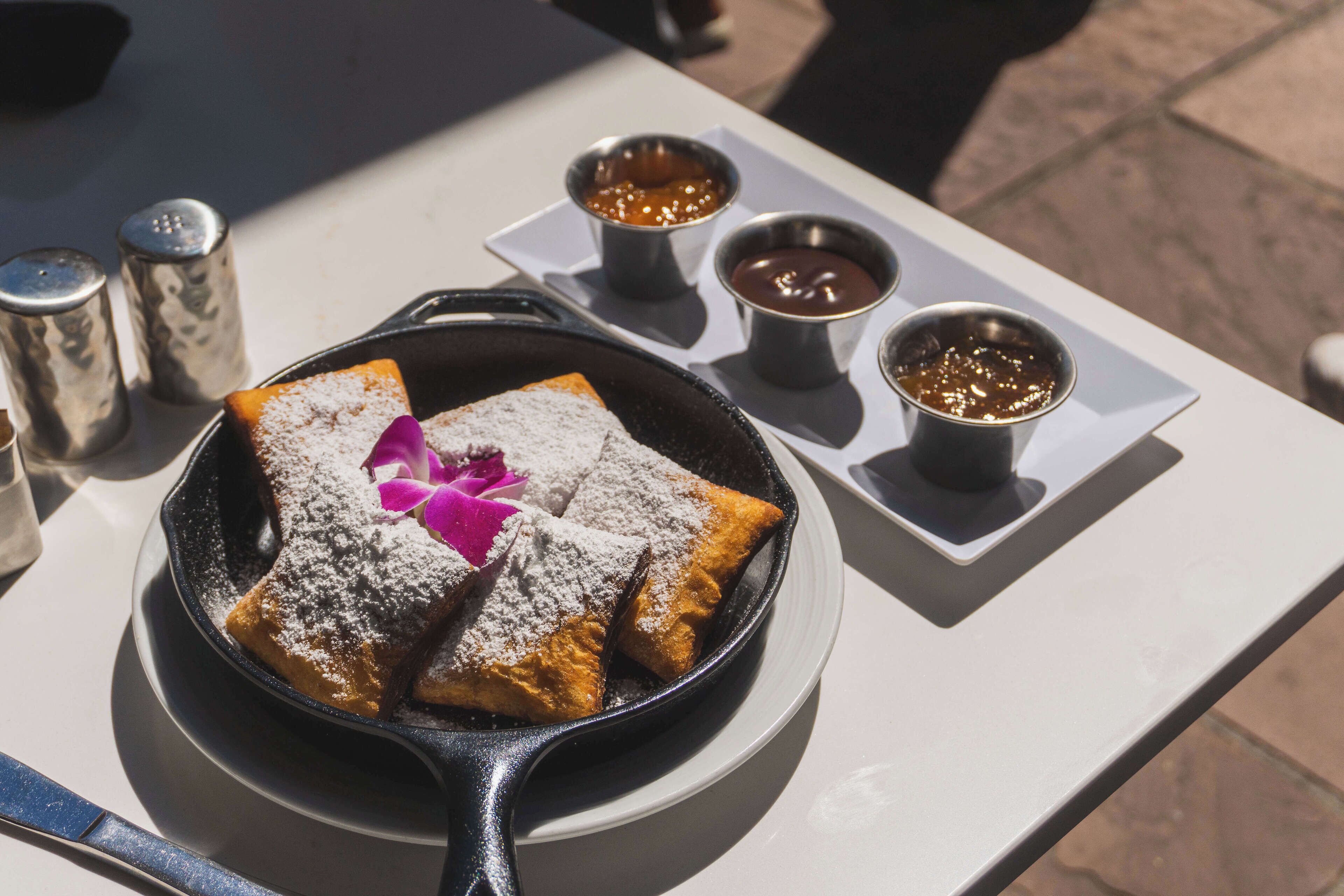 Ein Frühstückstisch mit einer Pfanne voller Pfannkuchen, einer rosa Blume und Puderzucker. Darauf stehen drei kleine Sirupfässer und zwei Salzstreuer.