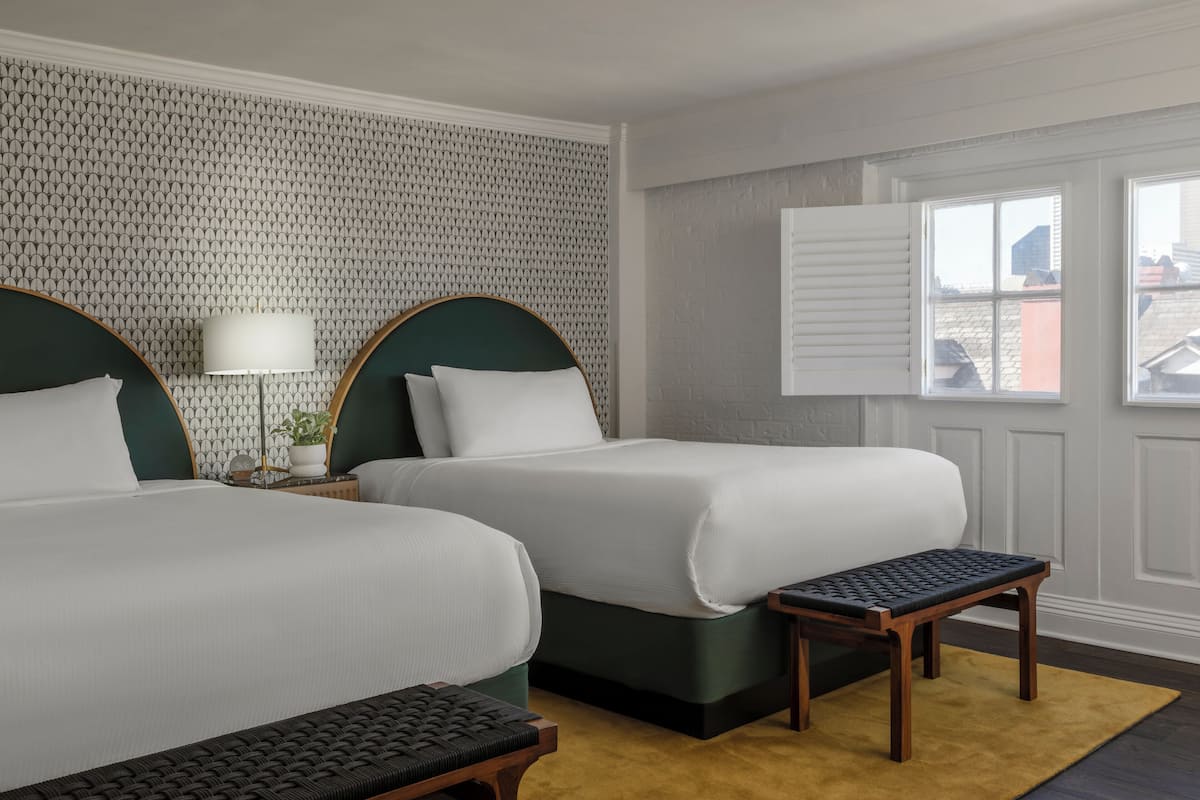 Een hotelkamer met twee bedden, een kleine tafel, een bankje en een raam met zonwering.
