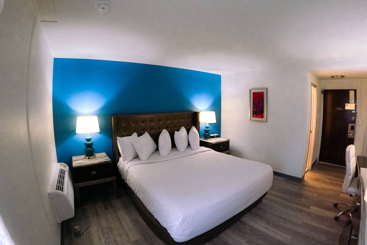 A hotel room with a large bed, two bedside tables with lamps, a wall-mounted air conditioner, and a door leading to another room.