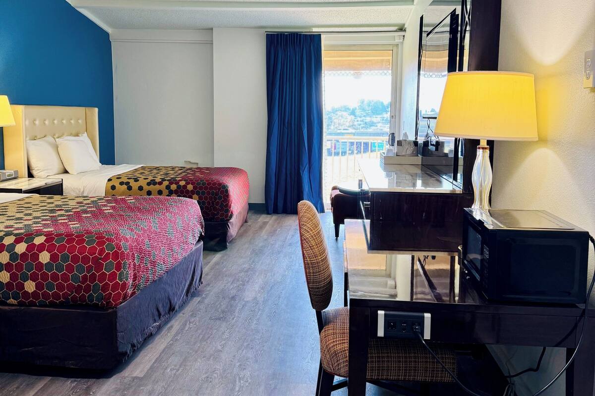 A hotel room with two beds, a desk, a chair, a lamp, a microwave, and a view of the outdoors.