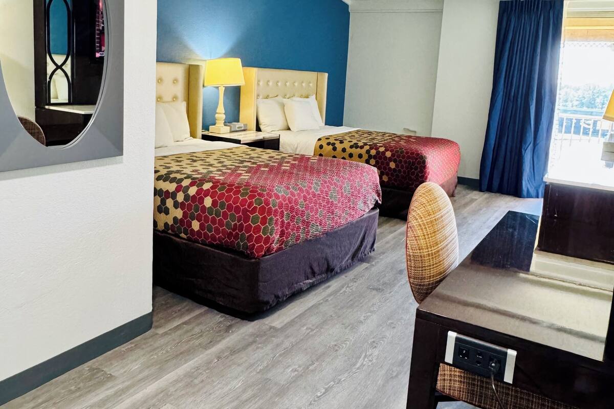 A hotel room with two beds, a round mirror, a desk, and a view of the outdoors.