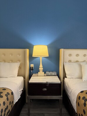 A hotel room with two beds, a nightstand with a lamp, a clock, and a power outlet.