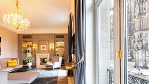 Un salón lujoso con candelabro, sofás, chimenea y vistas a un edificio histórico a través de un gran ventanal.