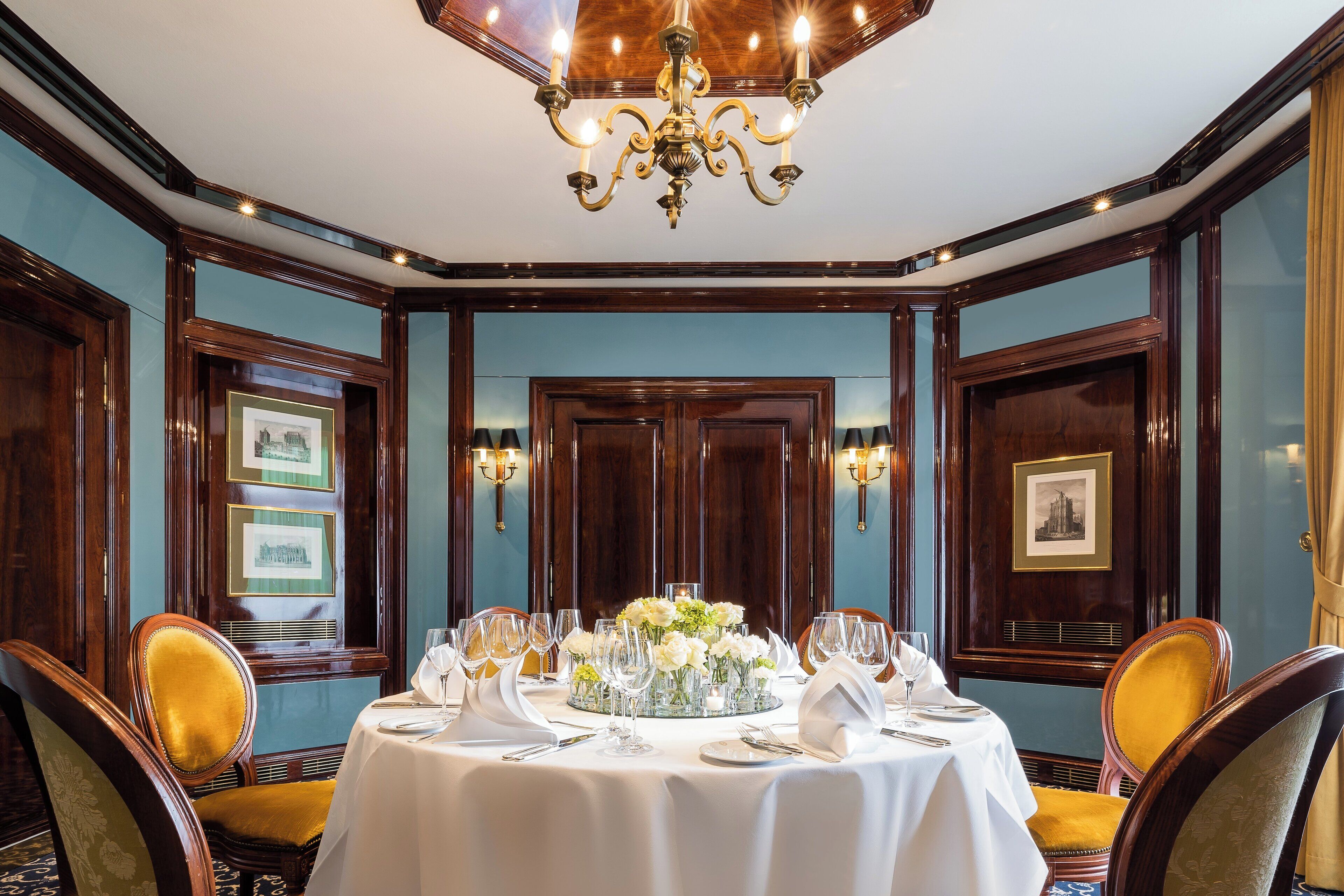 Une salle à manger élégante, avec une table ronde dressée pour six personnes, entourée de chaises richement décorées, d’un lustre et de tableaux encadrés accrochés aux murs.