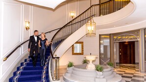 Una imponente escalera con alfombra azul y detalles dorados, que conduce a un espacio con piso de mármol, una fuente y plantas en macetas.