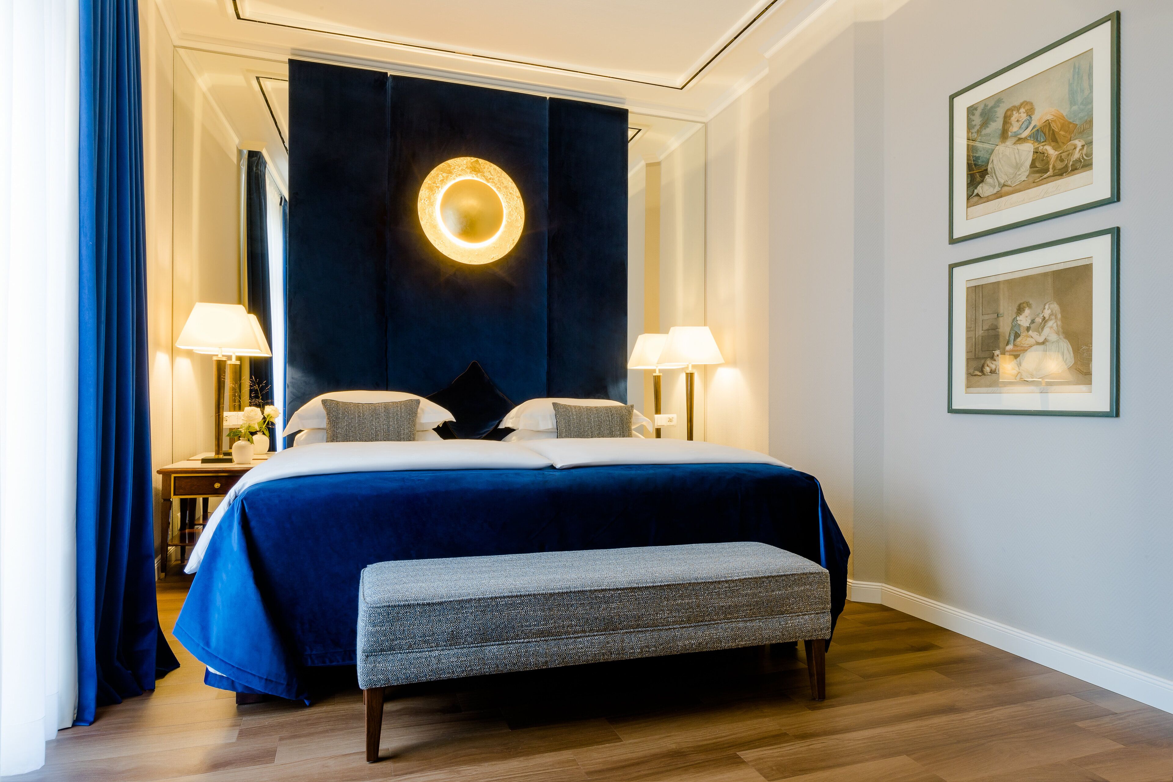 A hotel room with a large bed, two bedside lamps, a bench, and framed artwork on the wall.