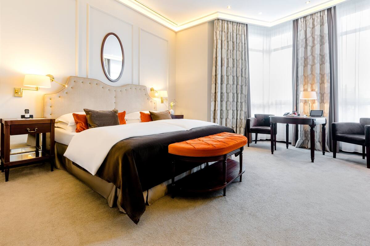 Quarto de hotel com uma cama grande, uma escrivaninha, duas cadeiras, um espelho e uma luminária.