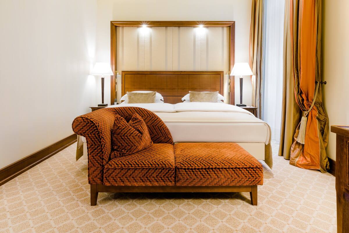 Quarto de hotel com uma cama, duas luminárias de cabeceira, uma poltrona estampada e uma cabeceira de madeira.