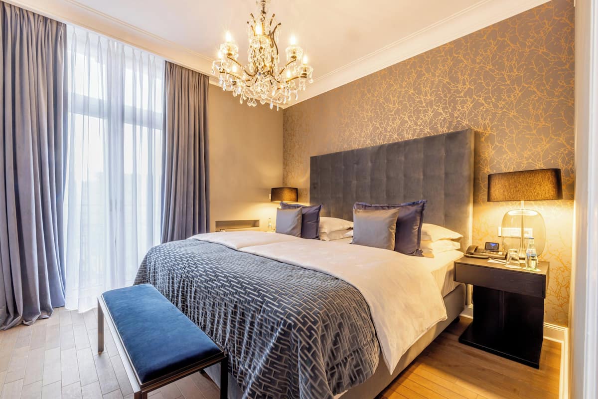 Quarto de hotel com uma cama grande, um banquinho, uma mesa de cabeceira, um lustre e parede estampada.