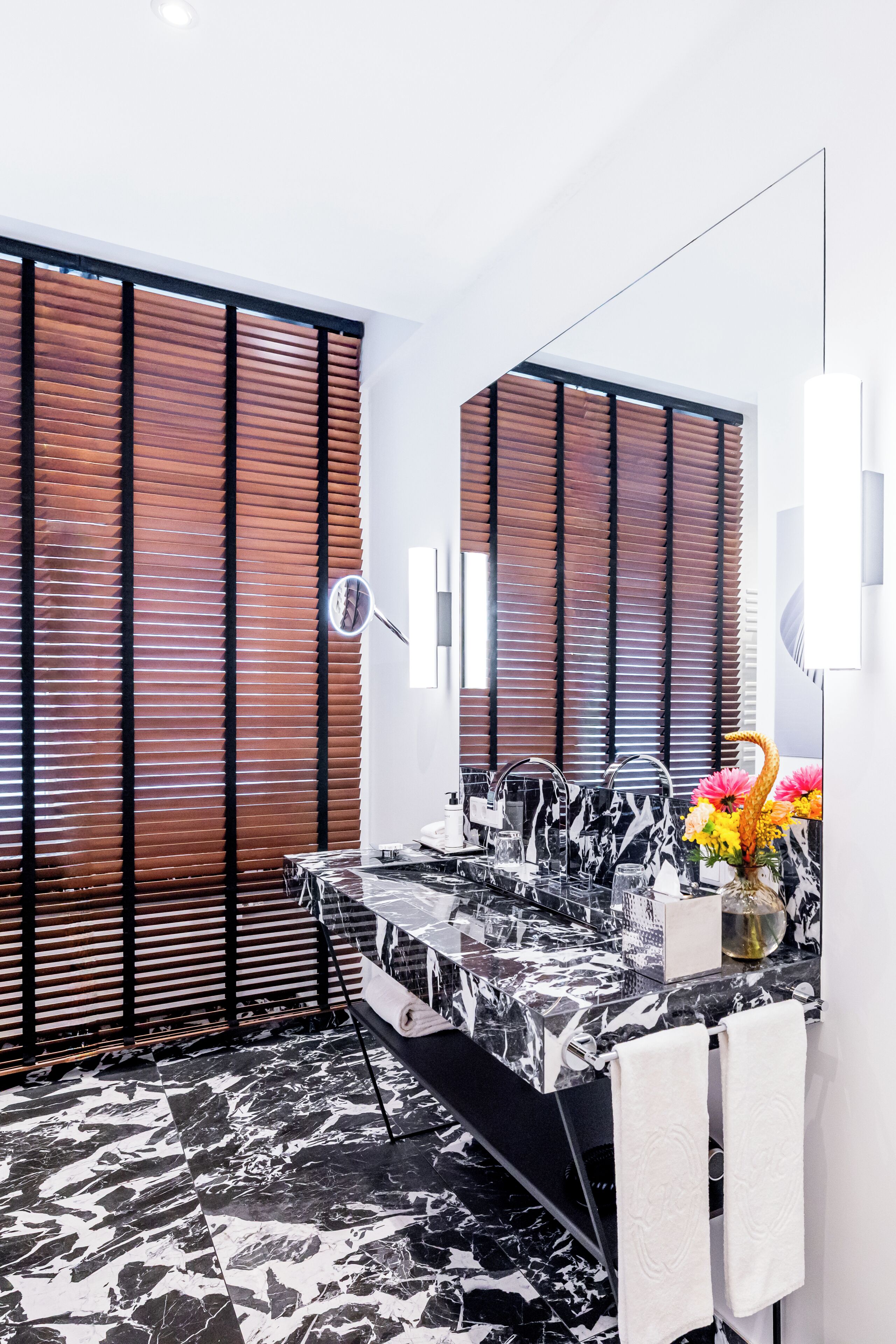 A bathroom with a black and white marble countertop, wooden blinds, a mirror, and a vase with flowers.