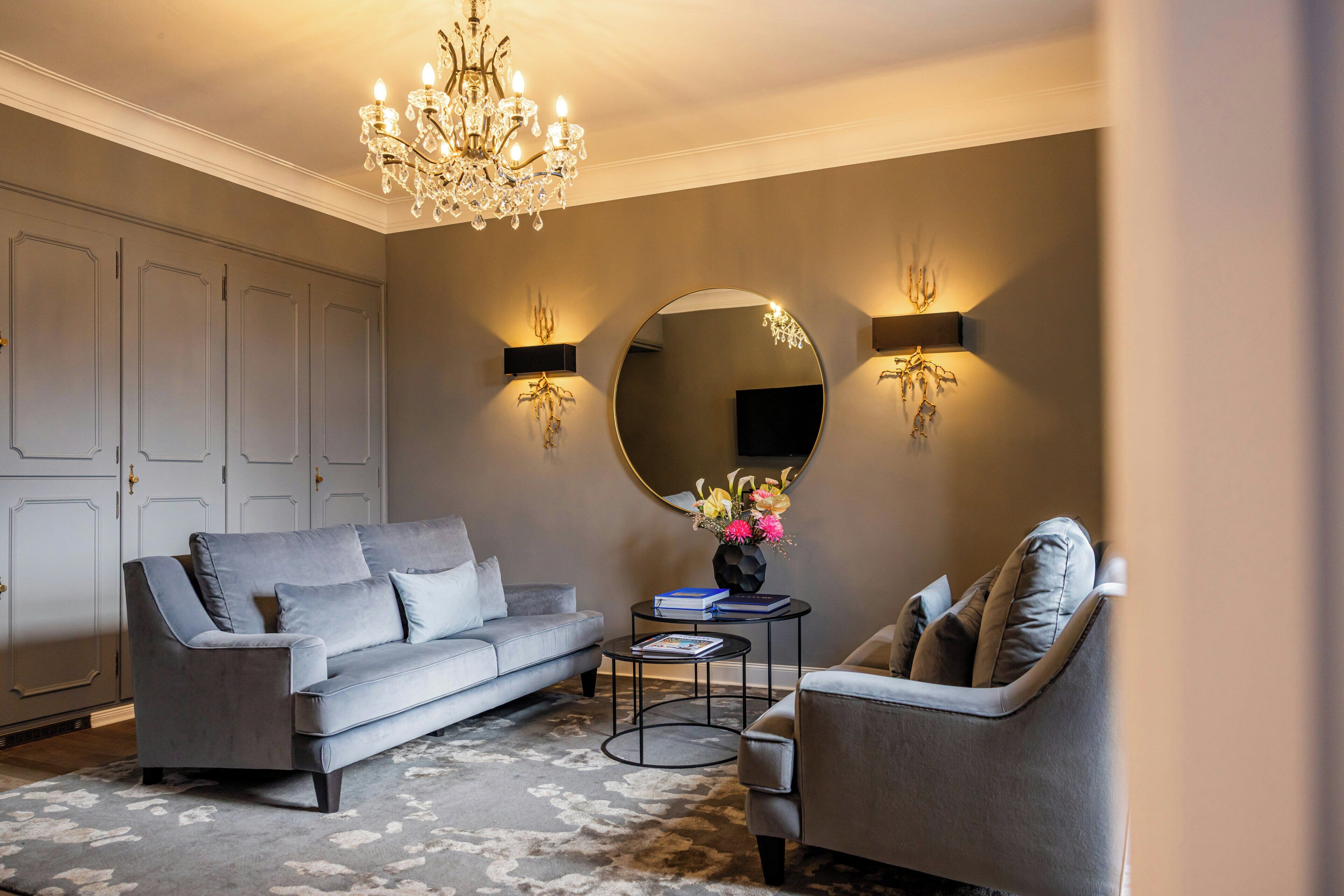 Un salon avec un canapé gris, un miroir rond, un lustre et deux lampes fixées au mur.