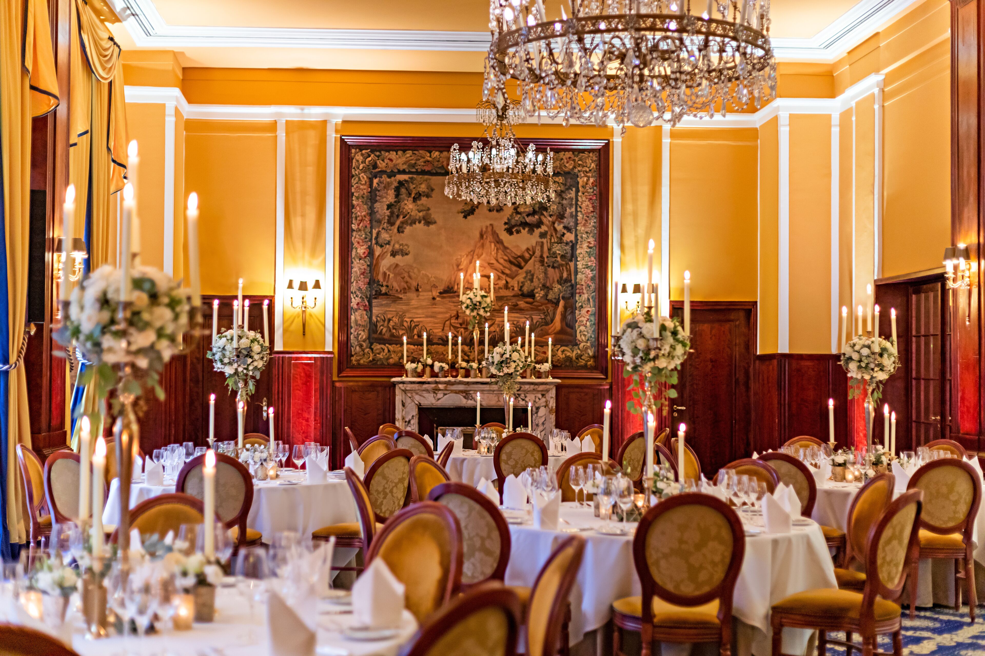 Ein formeller Speisesaal mit mehreren Tischen, gedeckt für ein Bankett, mit Kronleuchtern, Kerzen und einem großen Gemälde an der Wand.