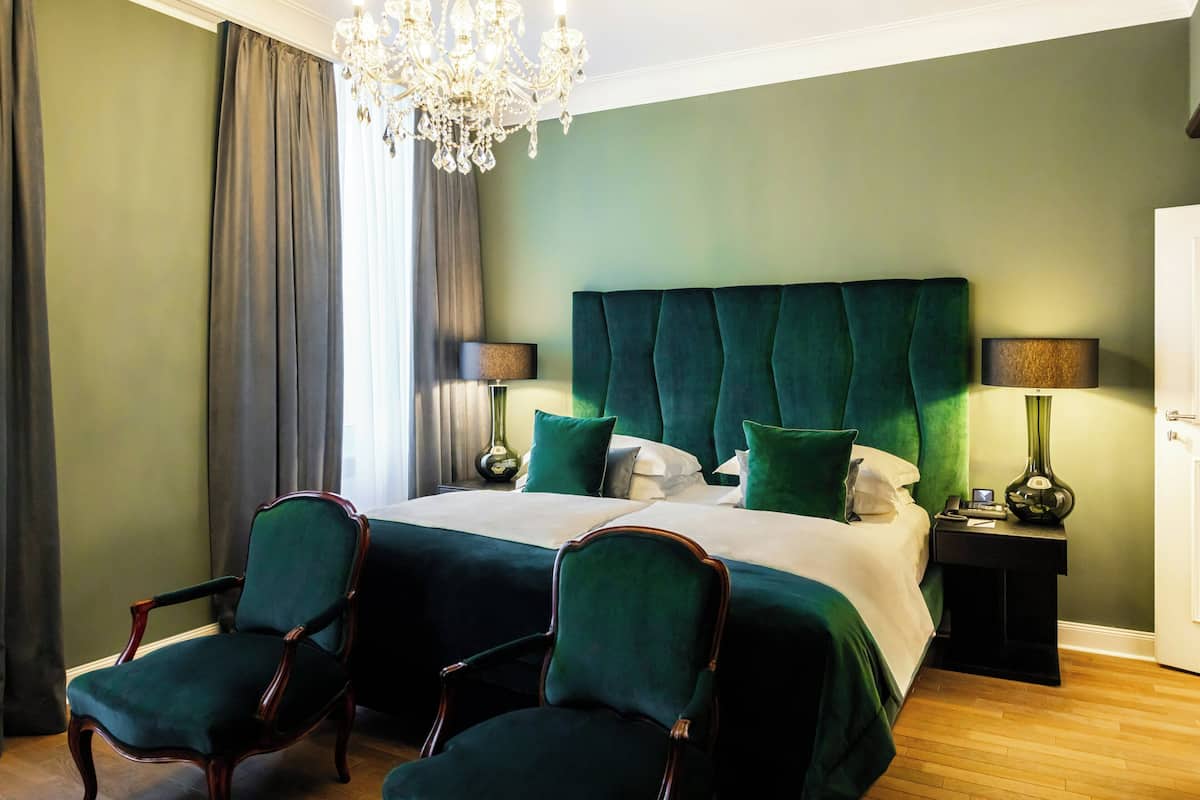 Uma cama de casal com roupa de cama verde, duas mesas de cabeceira, um lustre e duas poltronas estofadas em verde.