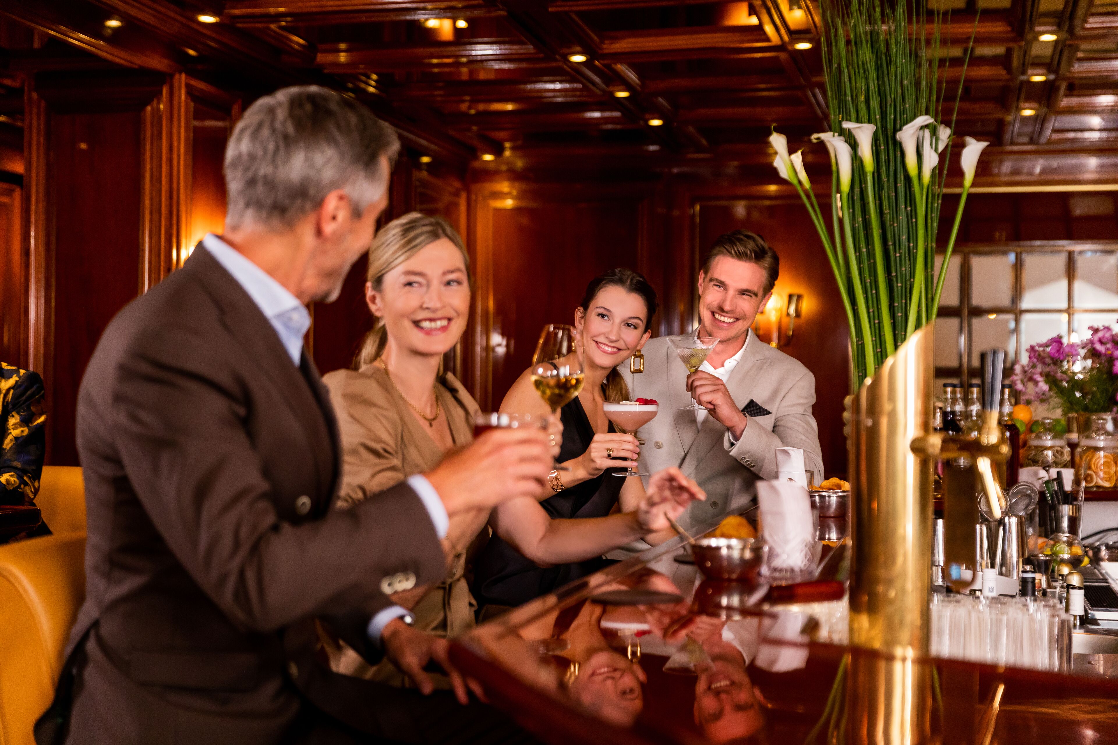 A group of people toasting at a bar with a variety of drinks and a vase of flowers.